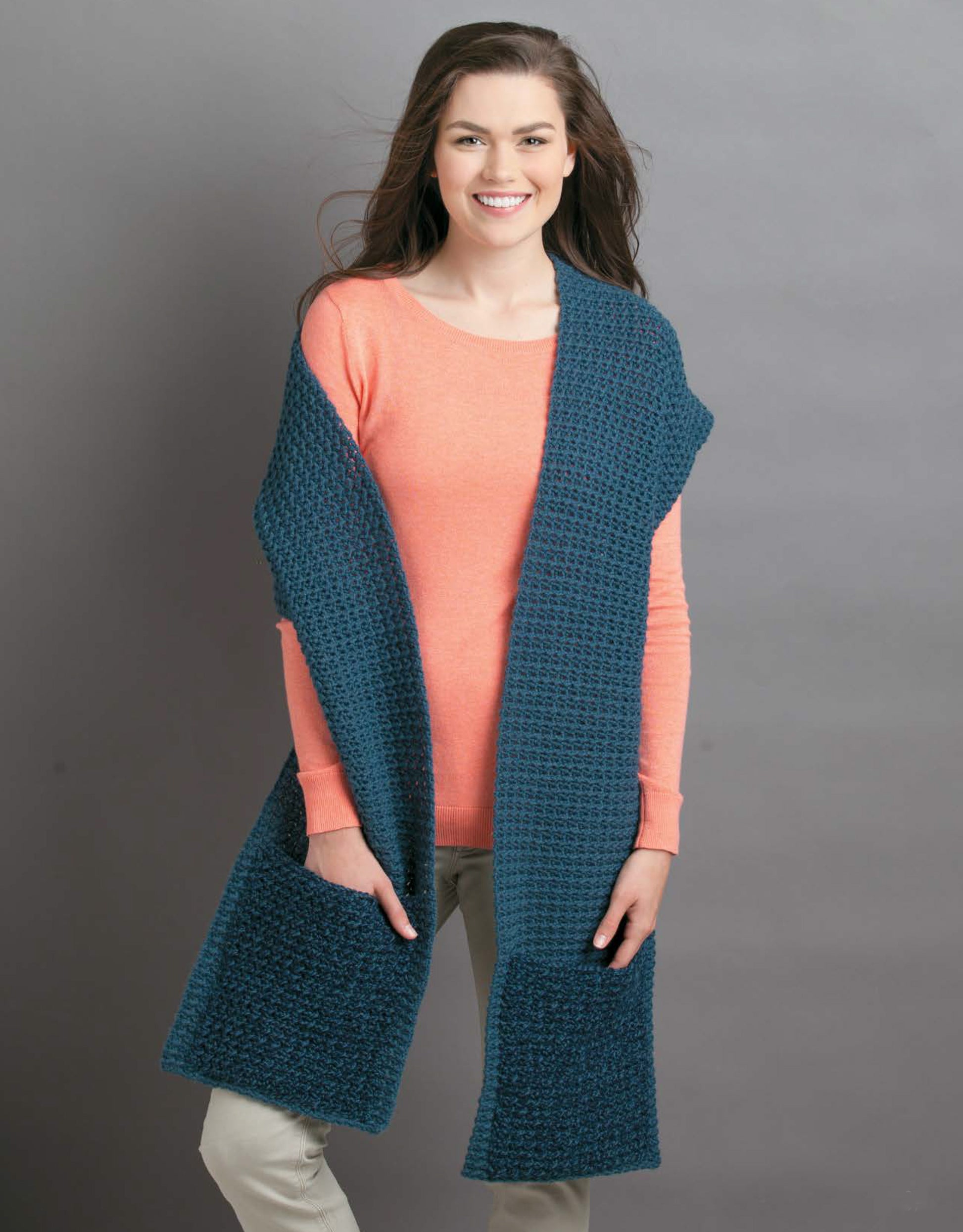 A woman with long brown hair models the Leisure Arts Pocket Shawls Digital Download, featuring a textured dark teal shawl with large pockets over a coral sweater and light pants, standing and smiling against a gray background.