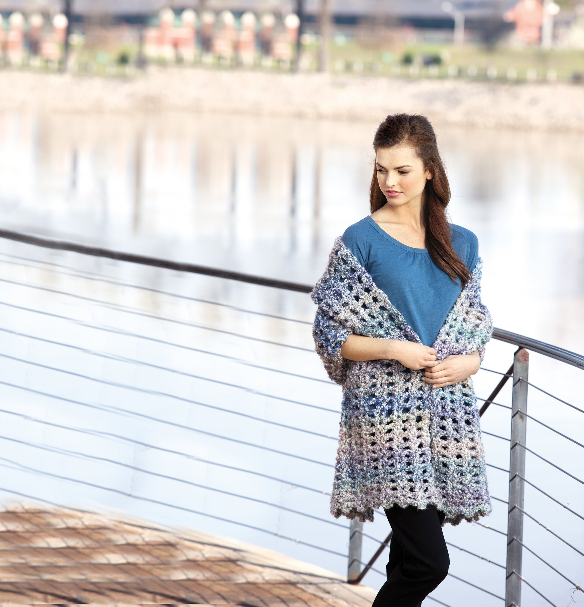 A woman stands by the water in a blue shirt, black pants, and a chunky multicolored "Healing Shawls" wrap from Leisure Arts—an inspiring look for anyone seeking unique crochet patterns or heartfelt handmade gifts.