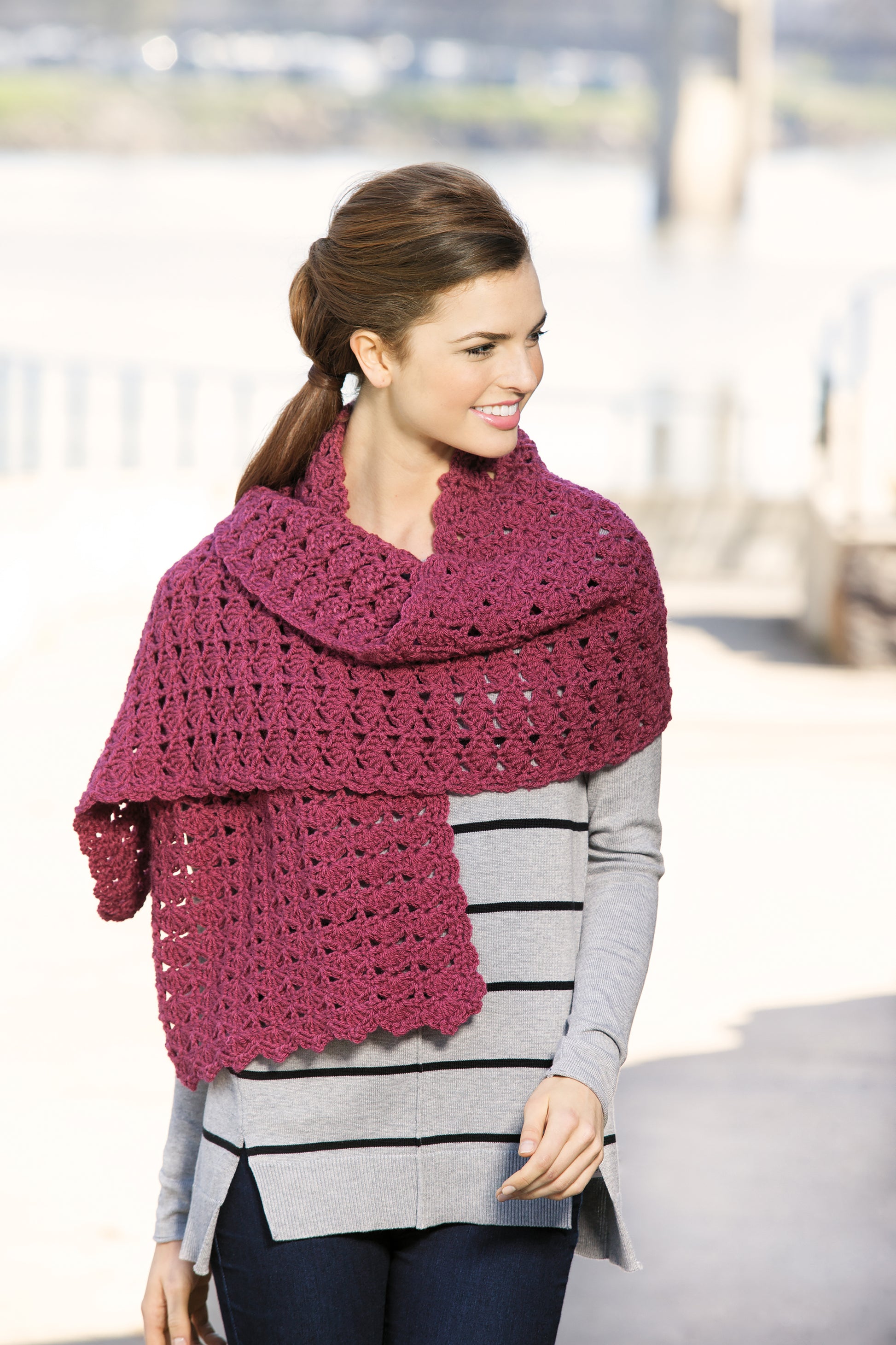 A woman with brown hair in a ponytail smiles outdoors, wearing Leisure Arts’ Healing Shawls—a large, textured dark pink crochet shawl. It’s perfect for unique gifts or stylish patterns. The bright background is softly blurred.