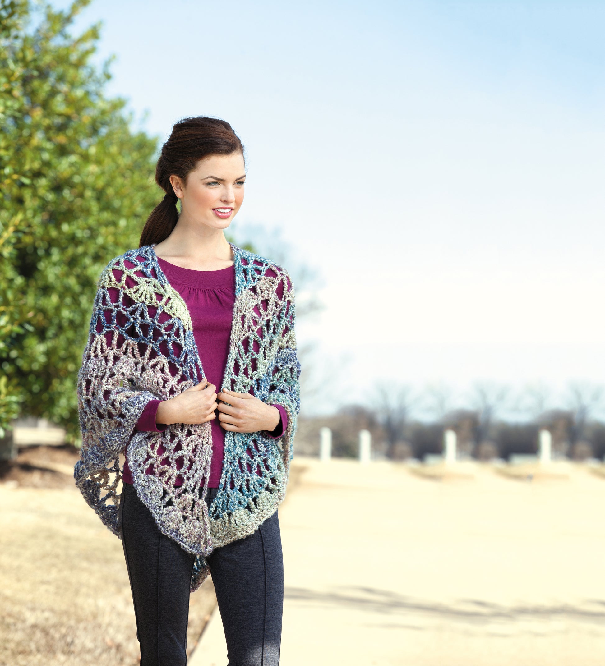 A woman stands outdoors on a sunny day, smiling as she wraps herself in the multicolored Healing Shawls by Leisure Arts, over a purple top and dark pants. Trees and blue sky form the background of this cozy crochet gift scene.