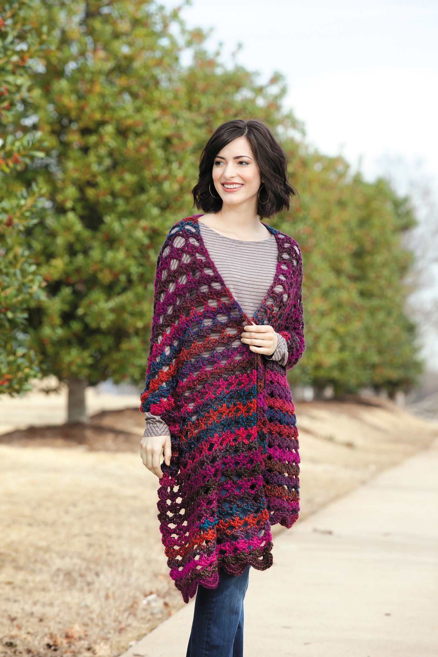 A woman with short dark hair smiles while wearing the Healing Shawls by Leisure Arts—a colorful, open-knit accessory inspired by crochet patterns—over a striped shirt and jeans, standing on a sidewalk beside green trees.