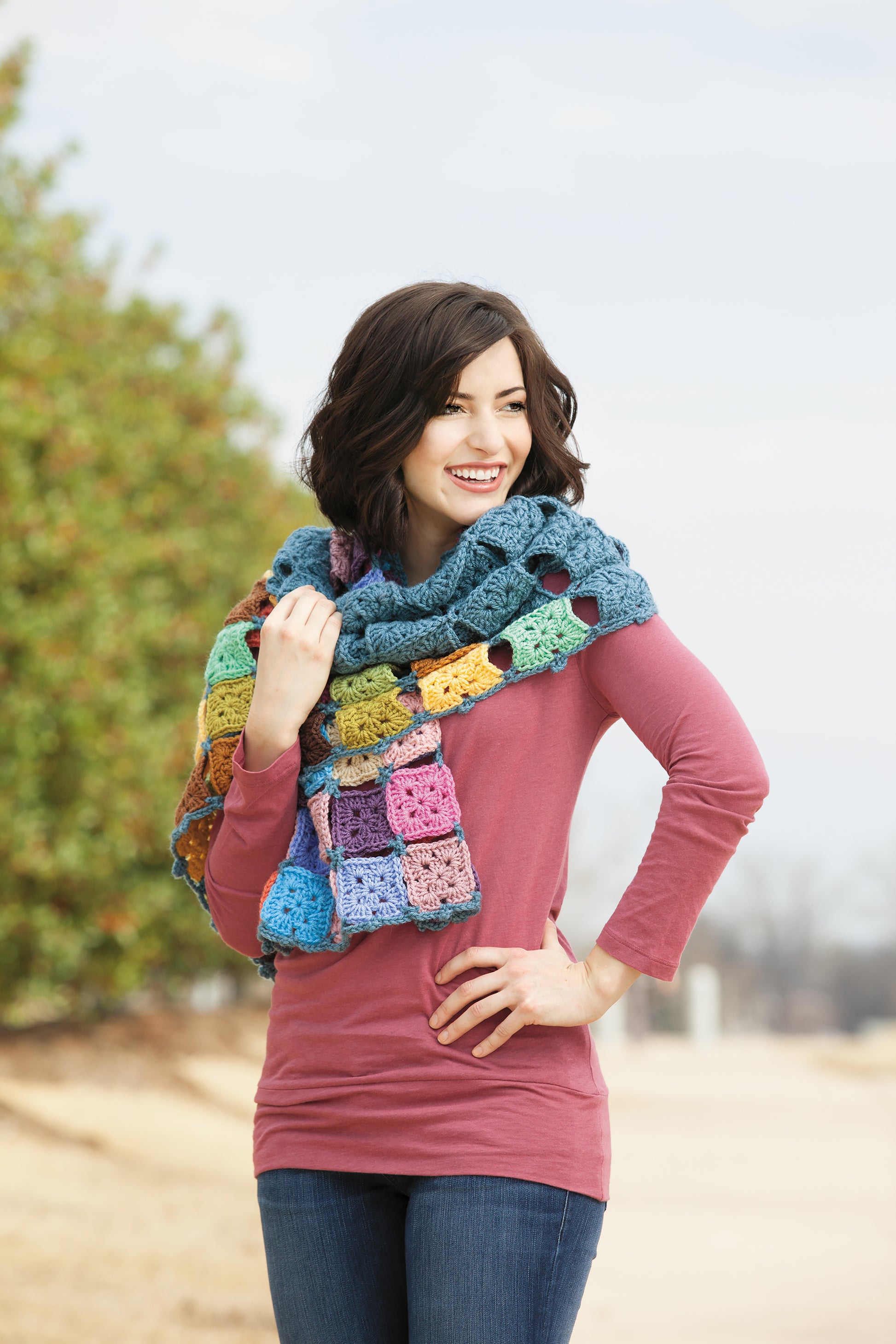 A smiling woman with short dark hair stands outside in jeans and a pink long-sleeve top, draped in a colorful Healing Shawl by Leisure Arts—an eye-catching crochet accessory. Trees and a blurred background add to the scene.