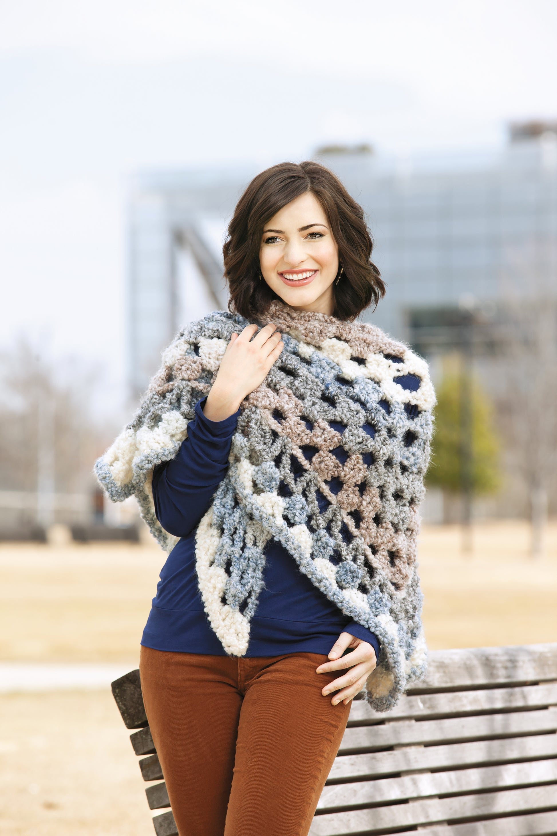 A woman with short dark hair stands outside, smiling as she wears the Leisure Arts Healing Shawls—a chunky, multicolored crochet piece—over a blue top and brown pants, one hand on a wooden bench. A modern glass building is behind her.