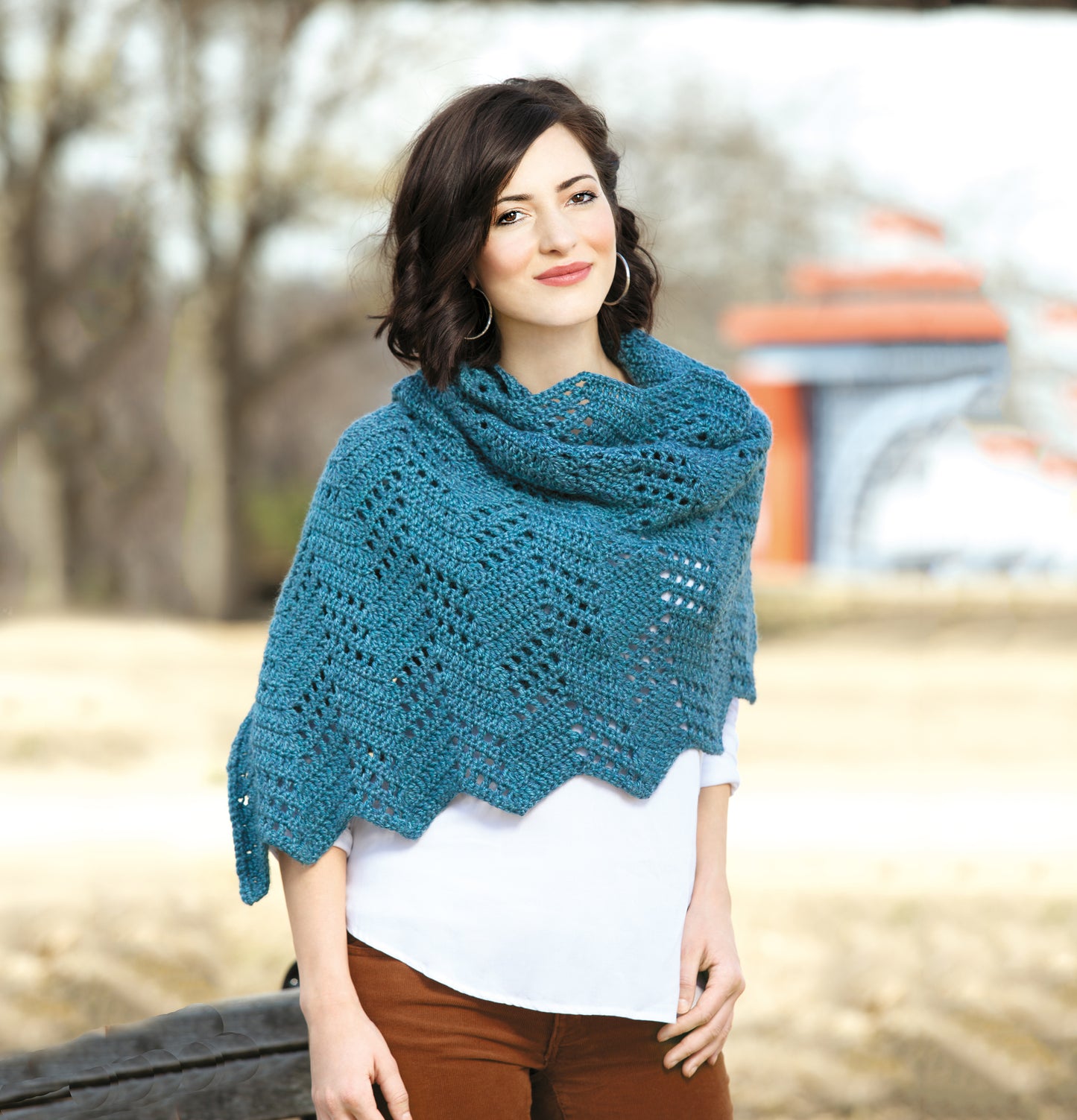 A woman with shoulder-length brown hair wears Leisure Arts’ Healing Shawls—a teal crochet accessory—over a white blouse and brown pants. This cozy piece is perfect for unique crochet gifts or anyone inspired by shawl patterns.