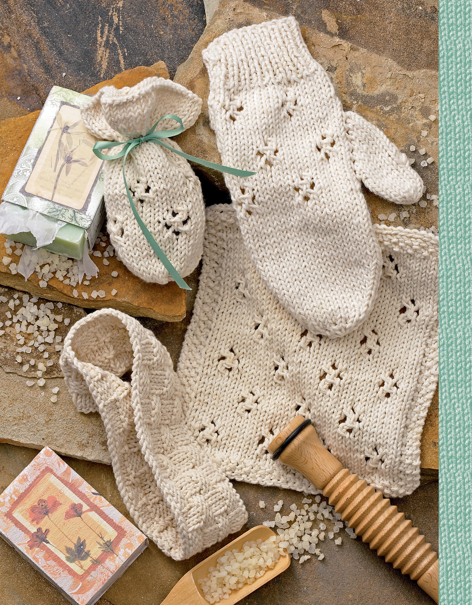 A flat lay showcases cream-colored hand-knits from Leisure Arts’ Spa & Bath Sets to Knit Digital Download—featuring a bath mitt, ribbon-tied pouch, headband, and washcloth with soap bars, grains, and a wooden massager on stone.