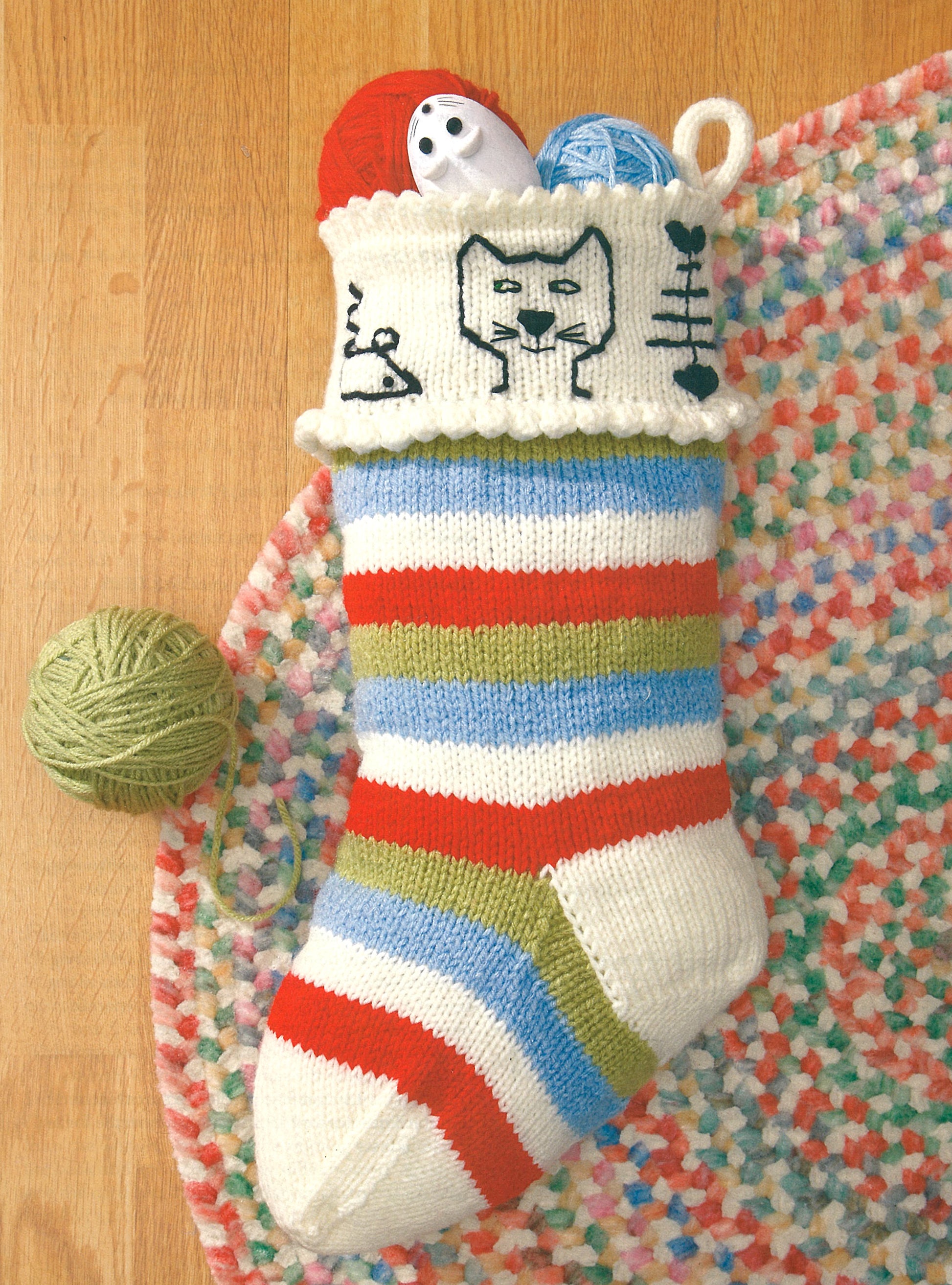 Leisure Arts' "The Stockings Were Knit" features a colorful striped Christmas stocking, ideal for intermediate crafters, filled with yarn balls and a snowman toy. The cuff is decorated with cat, fish, and mouse drawings.