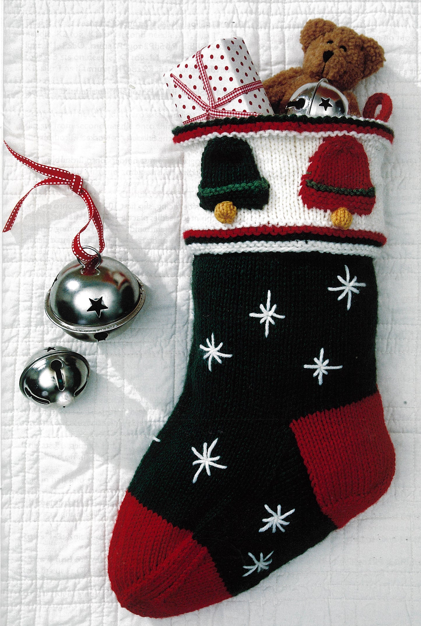 The Stockings Were Knit by Leisure Arts is an intermediate project featuring a green, red, and white Christmas stocking with star embroidery and bell motifs, stuffed with a teddy bear and gifts on a white quilted background.