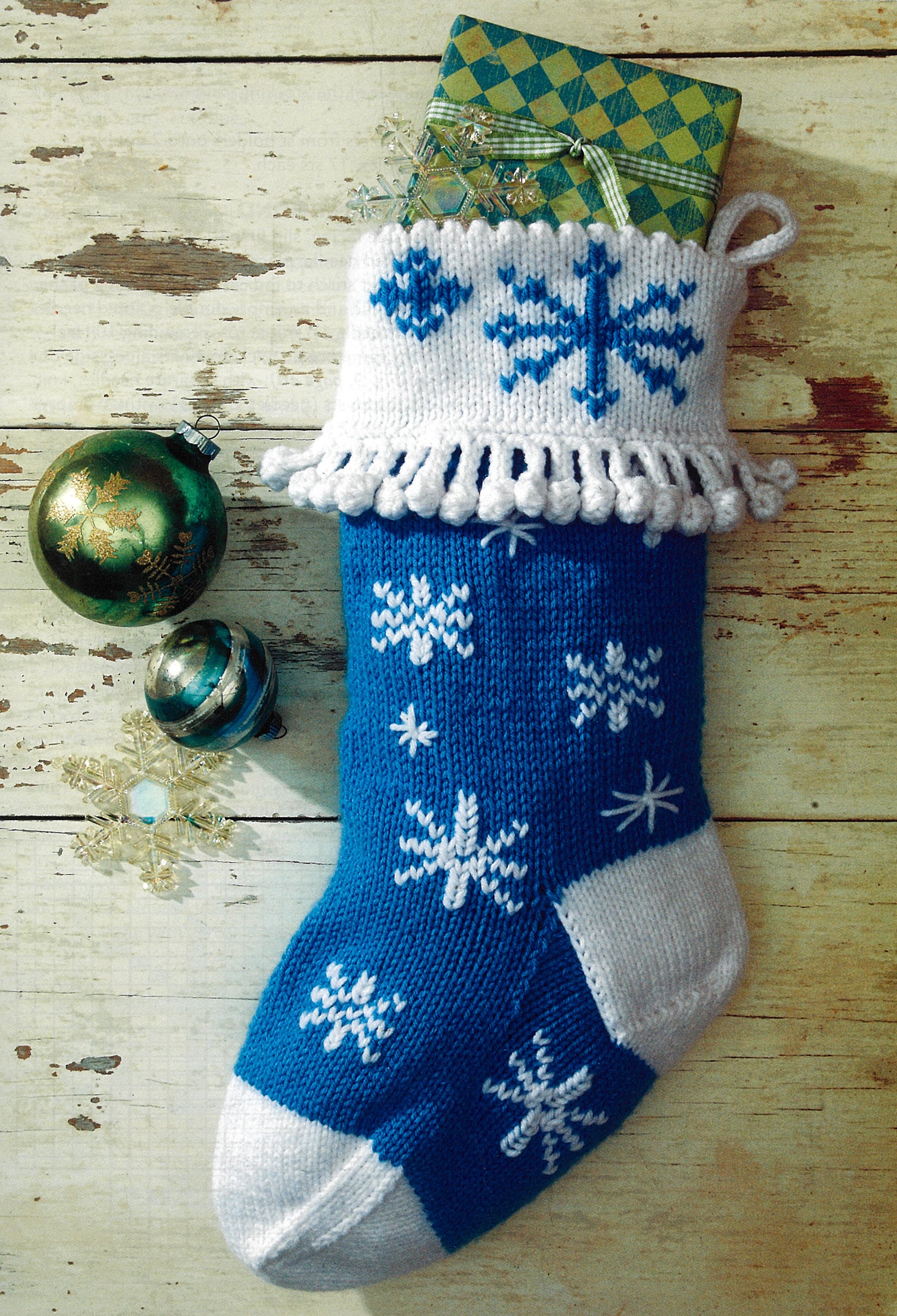 A blue and white Christmas stocking from Leisure Arts’ “The Stockings Were Knit” hangs on rustic wood with a gift inside. Green ornaments and clear snowflakes rest nearby—a memorable, intermediate knitting project.