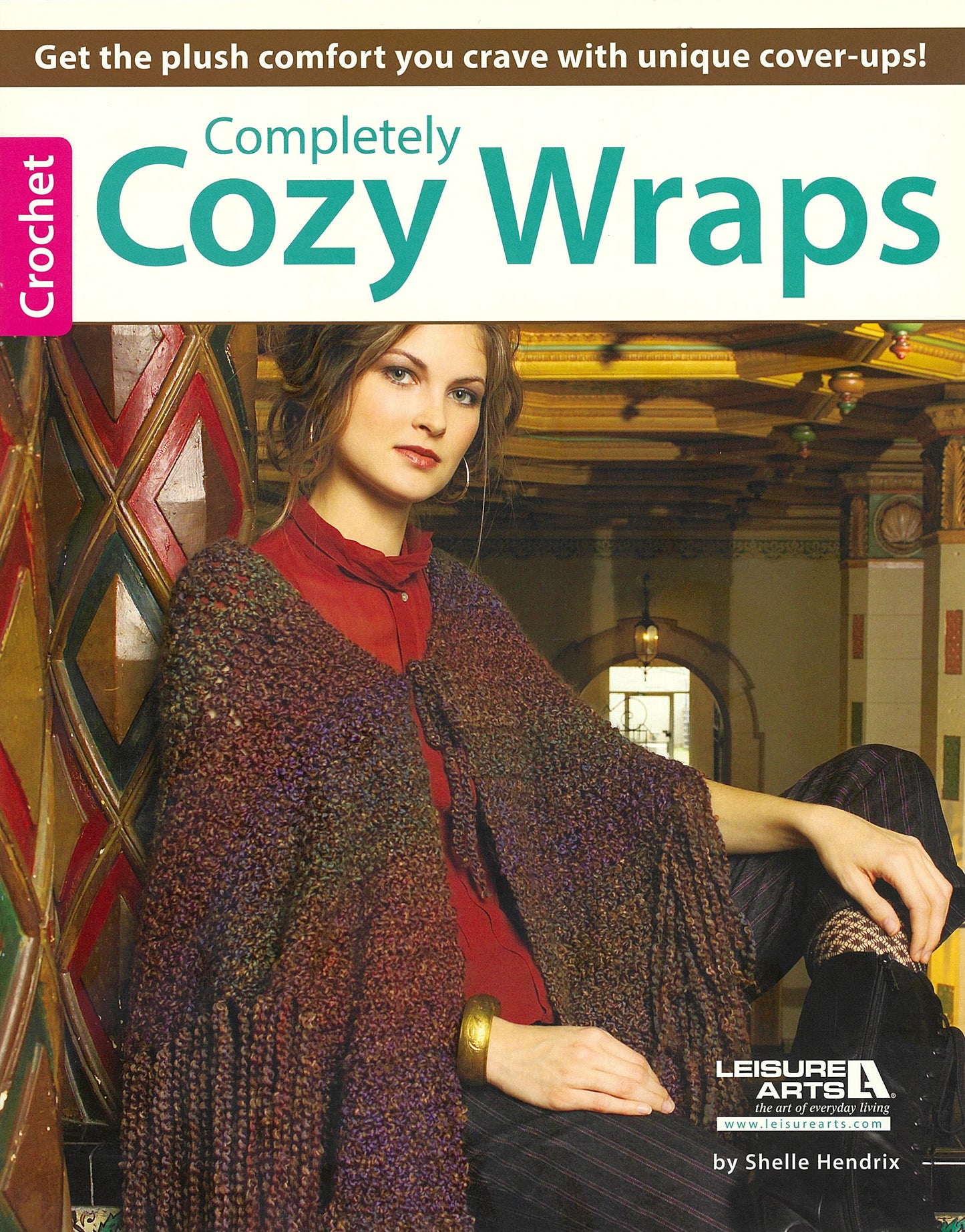 A woman in a chunky, multicolored crocheted wrap sits on stairs in a vintage space. The Leisure Arts "Completely Cozy Wraps" magazine showcases unique wraps for plush comfort and style.