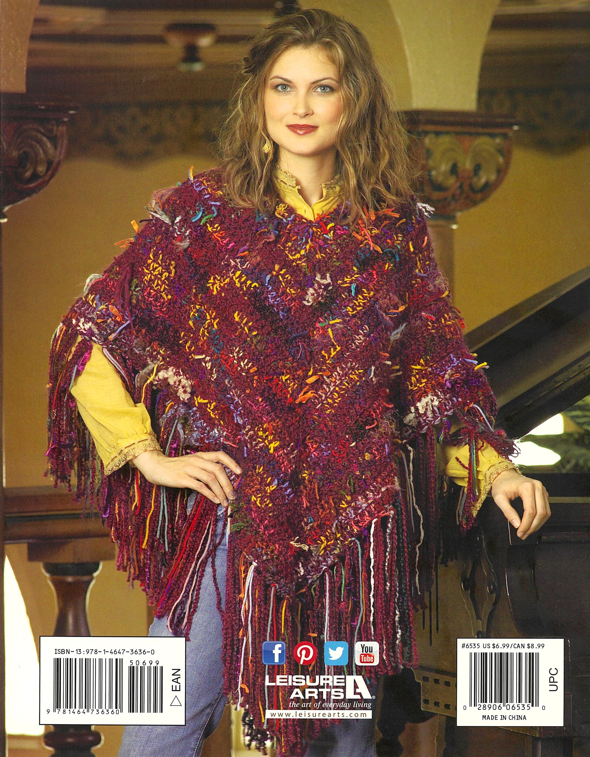 A woman with wavy brown hair wears a Completely Cozy Wrap by Leisure Arts over a yellow shirt. Indoors, she smiles softly with one hand on a piano, framed by ornate columns and arches that complement her stylish comfort.