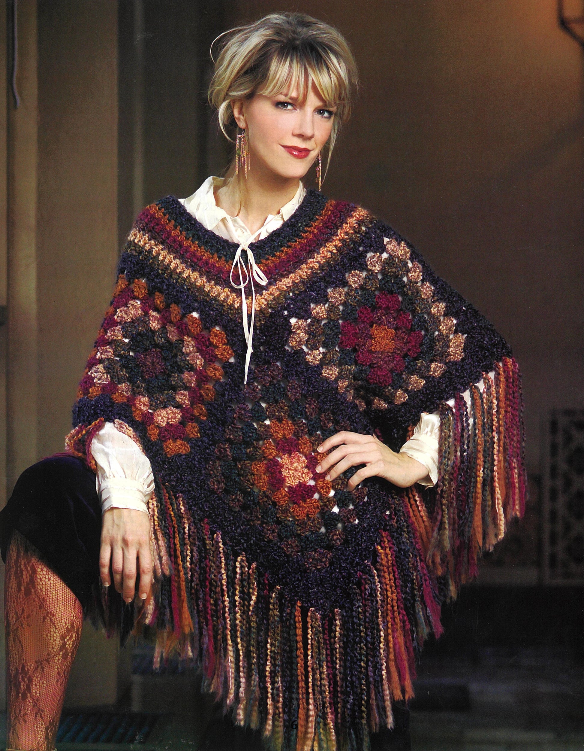 A blonde woman models the Leisure Arts "Completely Cozy Wraps" poncho, featuring colorful granny squares and long fringe, over a tie-neck white blouse. She poses indoors with one hand on her knee.