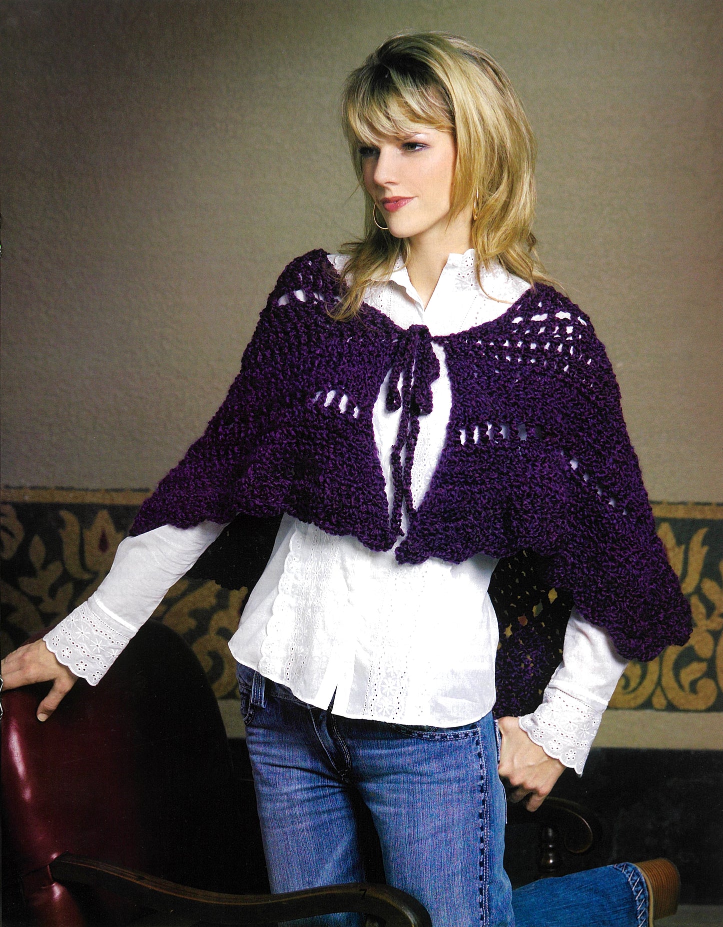A woman in a white blouse, blue jeans, and a chunky purple shawl from Leisure Arts' Completely Cozy Wraps stands indoors by a dark red chair with a patterned wall behind her, radiating plush comfort.