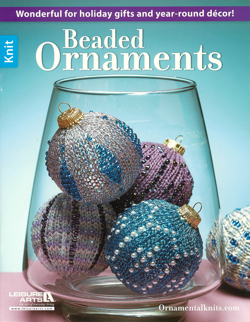 Four beaded, knitted ornaments in blue, purple, and silver rest inside a clear glass vase. The Leisure Arts Beaded Ornaments magazine showcases holiday gifts and year-round decor.