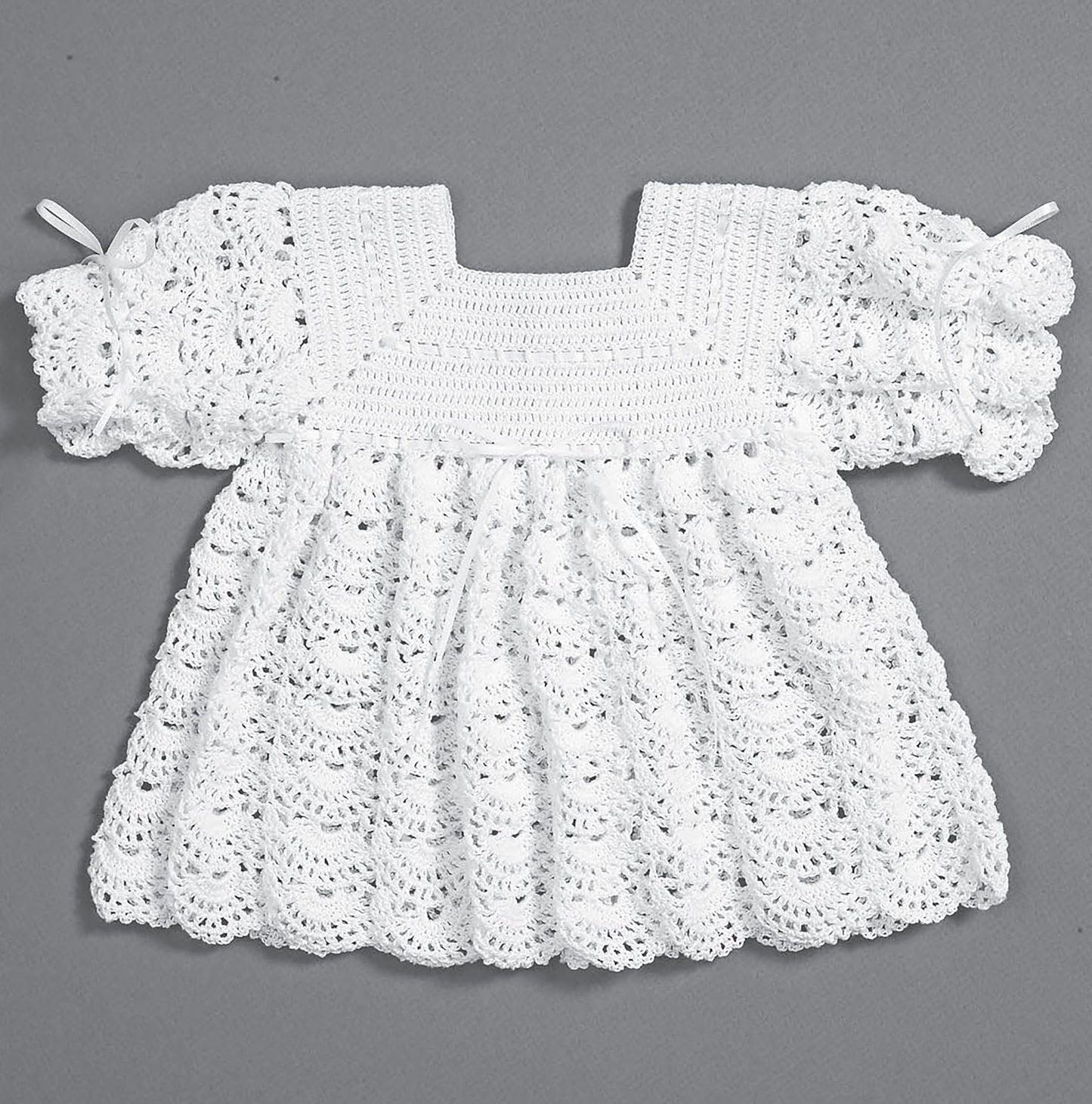 The Christening Sets by Leisure Arts—an heirloom white hand-crocheted baby dress with short puffy sleeves and a scalloped lacy pattern—is shown flat against a gray background. Celebrate your newborn with this adorable outfit!.