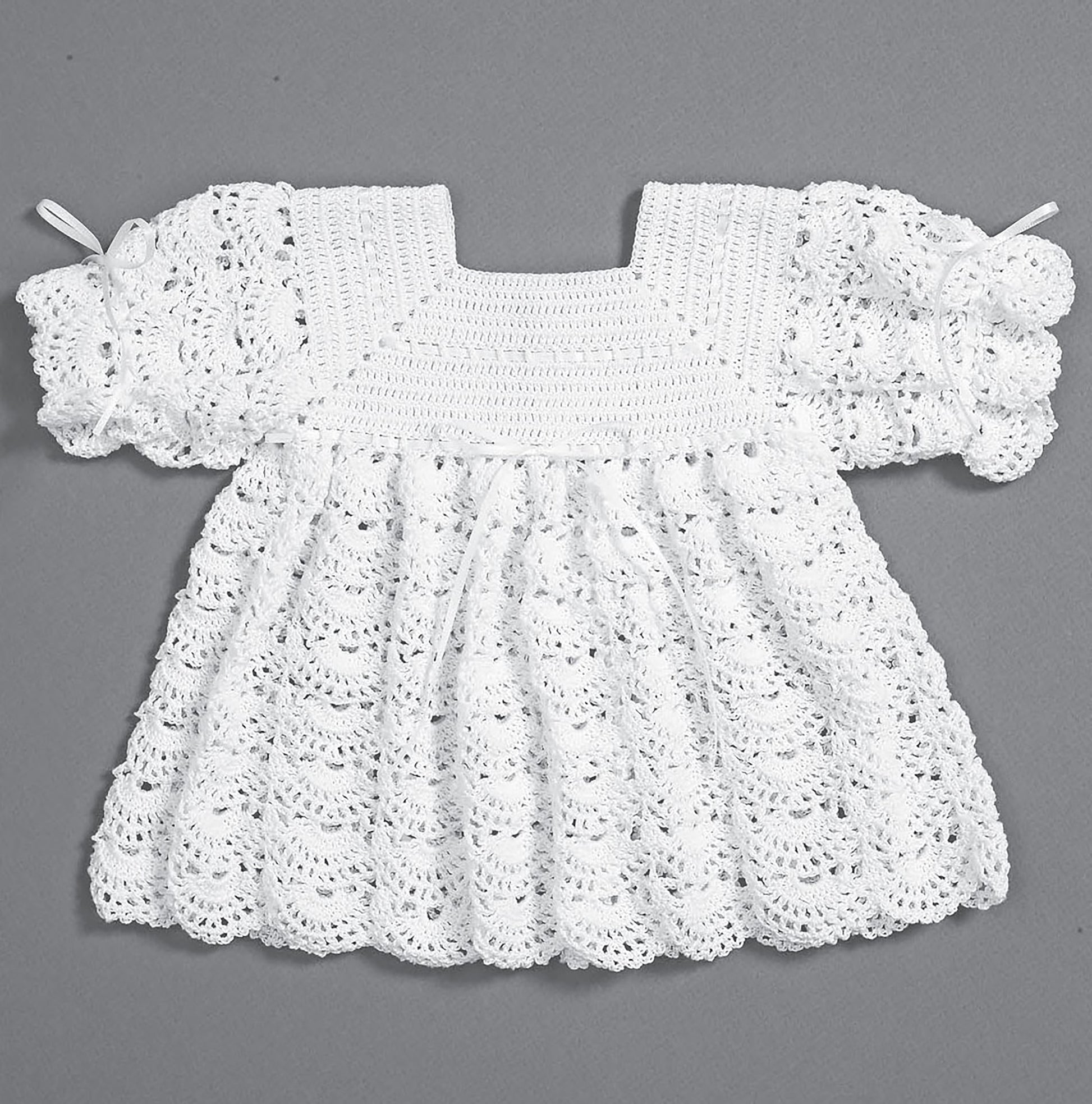 The Christening Sets by Leisure Arts—an heirloom white hand-crocheted baby dress with short puffy sleeves and a scalloped lacy pattern—is shown flat against a gray background. Celebrate your newborn with this adorable outfit!.