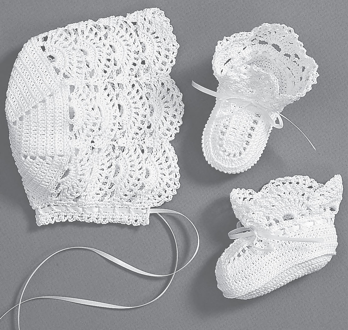 The Leisure Arts Christening Sets—Celebrate Your Newborn With an Heirloom Outfit! features a delicate white crochet baby bonnet with ribbon ties and matching booties with bows and lace patterns, displayed on a gray background.
