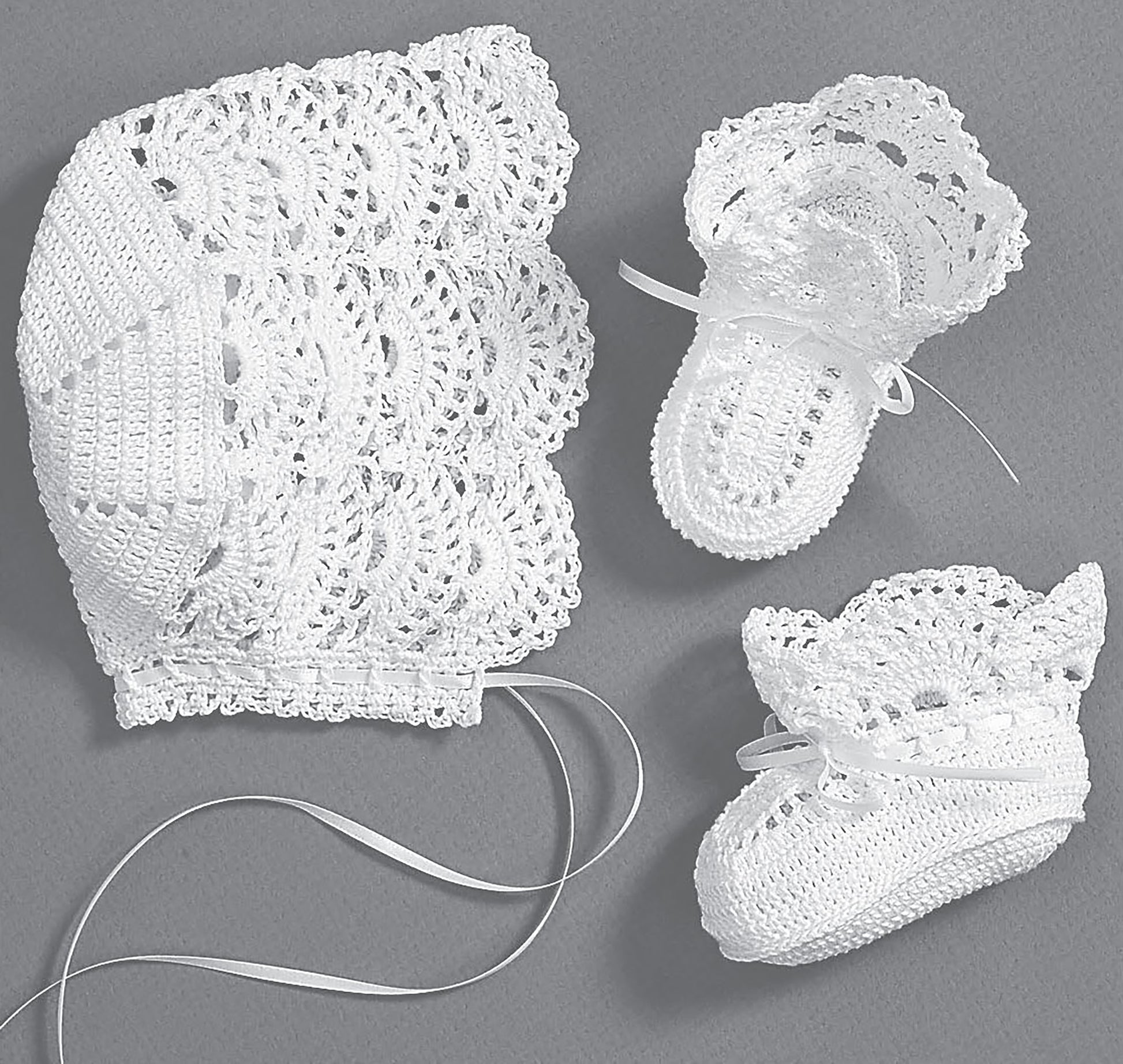 The Leisure Arts Christening Sets—Celebrate Your Newborn With an Heirloom Outfit! features a delicate white crochet baby bonnet with ribbon ties and matching booties with bows and lace patterns, displayed on a gray background.