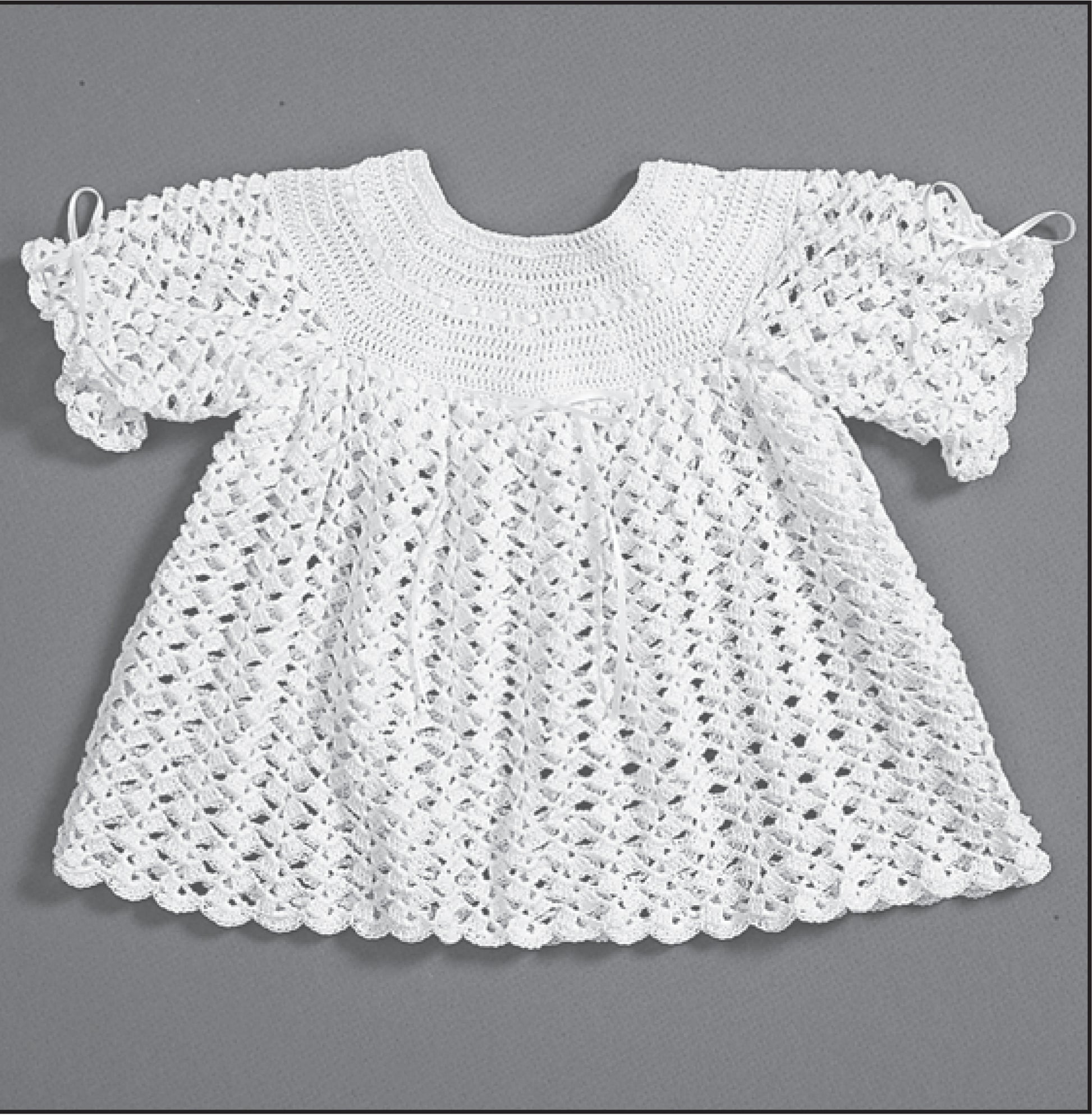 Leisure Arts’ Christening Sets—Celebrate Your Newborn With an Heirloom Outfit! This white, short-sleeved, crocheted baby dress features a lacy openwork pattern, textured top, and scalloped edges. Displayed flat on a gray background.