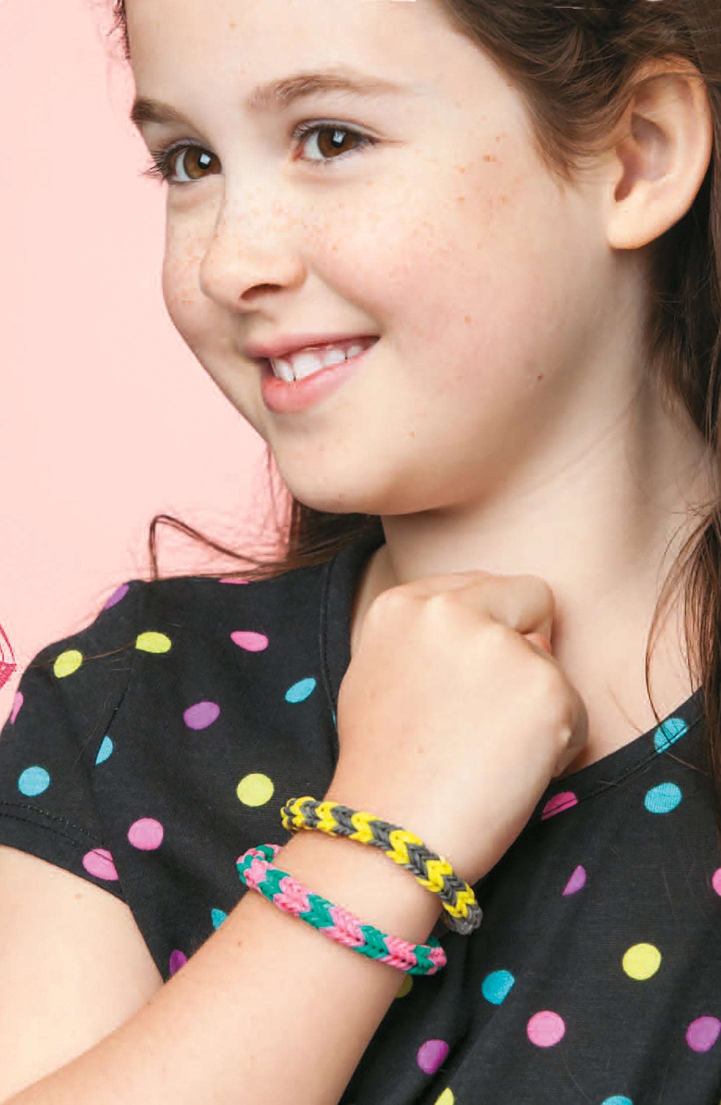 A smiling girl with long brown hair, in a black shirt with colorful polka dots, proudly displays two DIY braided bracelets made using Monster Tail Rubber Band Projects by Leisure Arts—perfect for step-by-step bracelet designs against a pink backdrop.