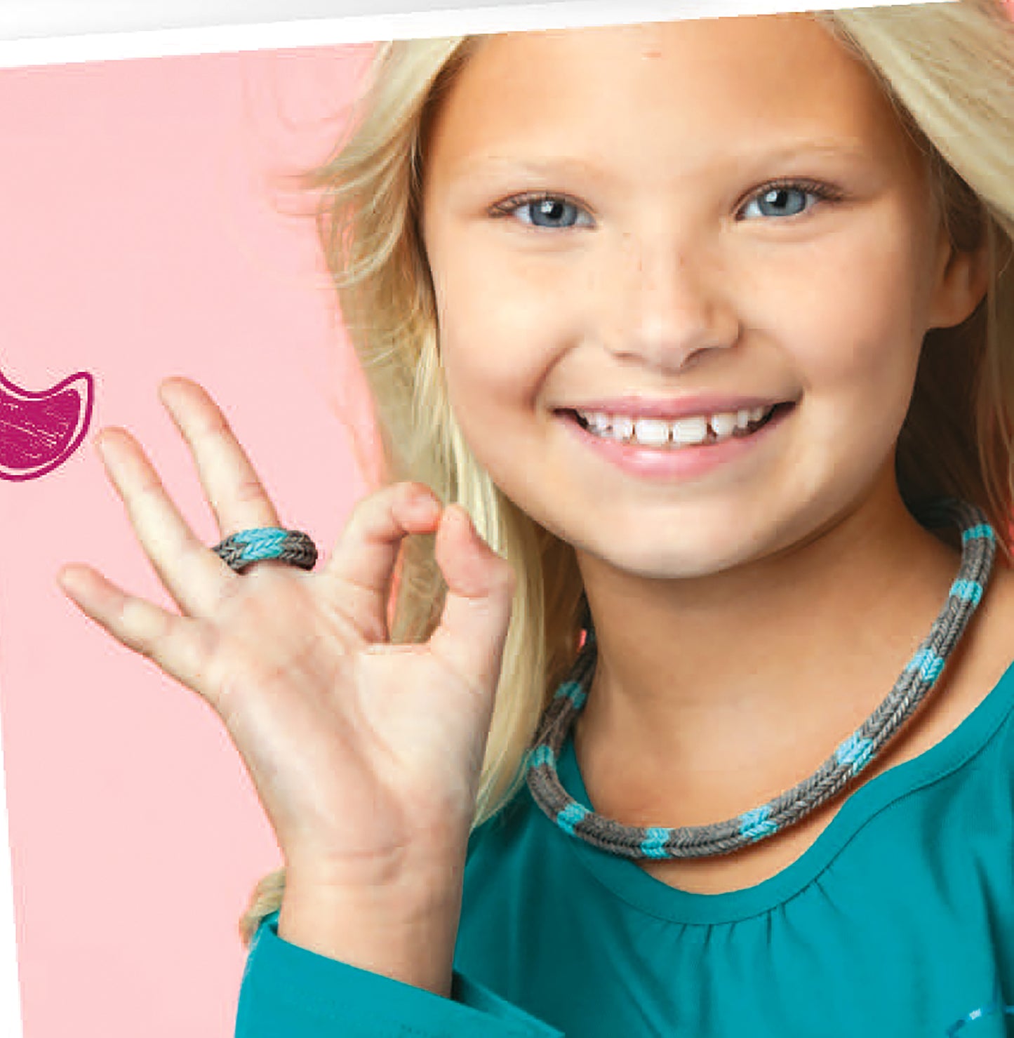A smiling young girl with long blonde hair in a teal shirt shows an OK gesture, ready for bracelet making fun with Leisure Arts’ Monster Tail Rubber Band Projects—perfect for kids who love jewelry crafts.