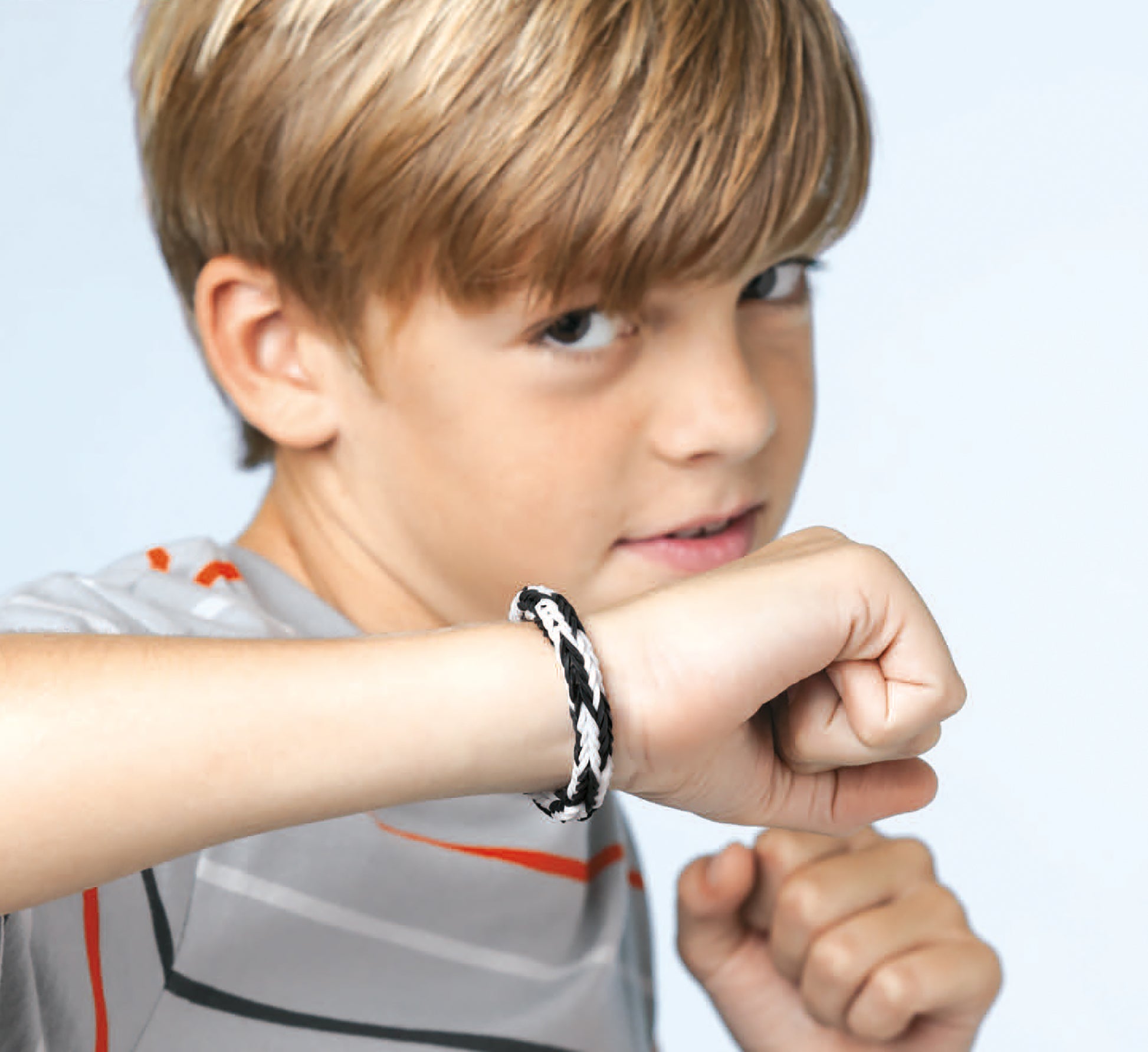 A young blond boy in a gray shirt wears a black-and-white braided bracelet made with Monster Tail Rubber Band Projects from Leisure Arts, showing off his wrist and looking confidently at the camera.