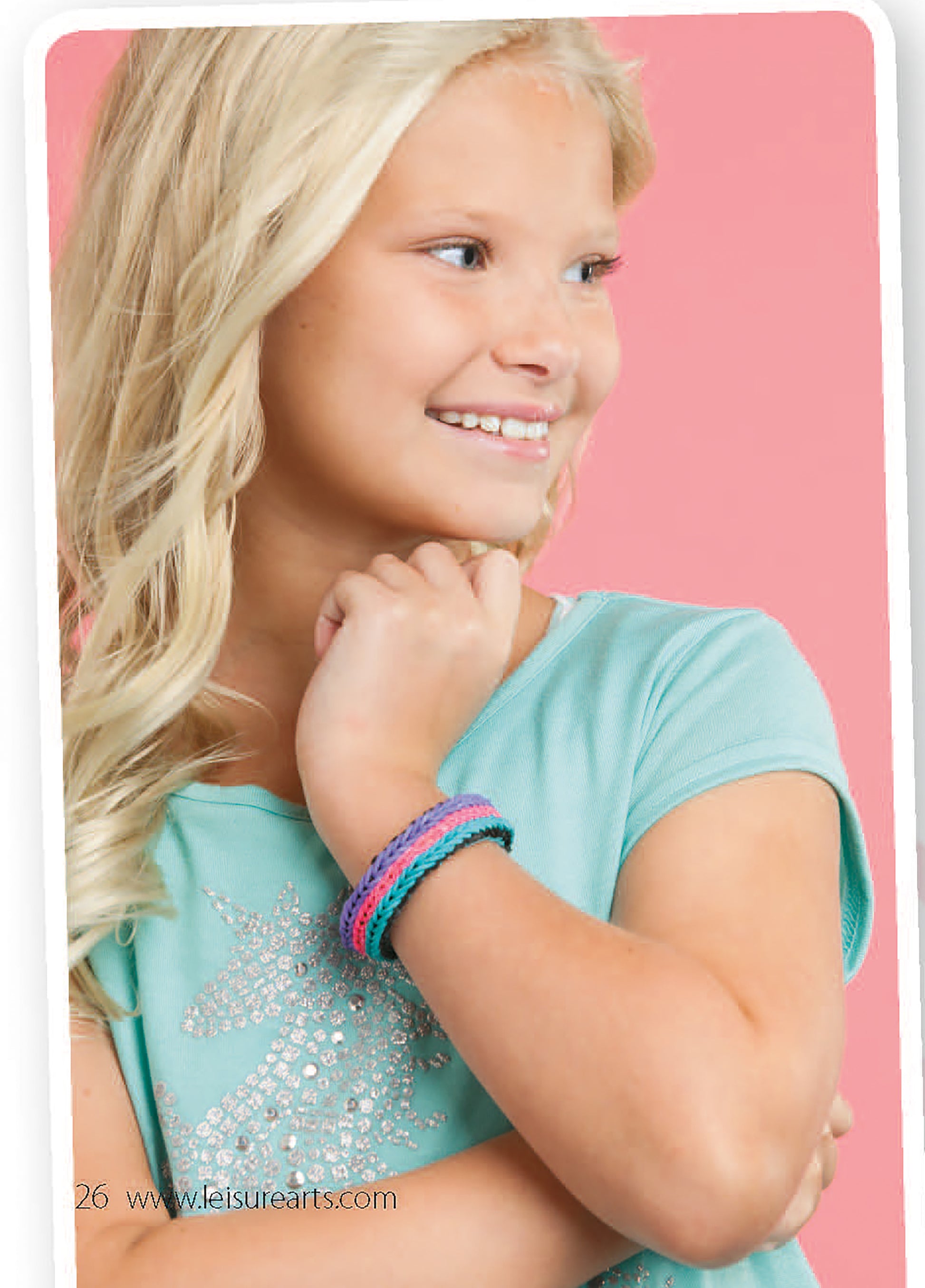 A smiling young girl with long blond hair wears a light blue shirt with a sparkling unicorn, posing against a pink background and showing off braided bracelets she made herself using the Monster Tail Rubber Band Projects from Leisure Arts.