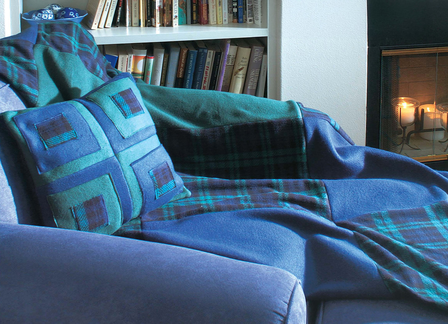 A cozy living room with a blue and green Fleece at Home blanket from Leisure Arts, a matching cushion on a blue armchair, books on a white shelf, and candles in the fireplace—perfect for relaxing with easy throws.