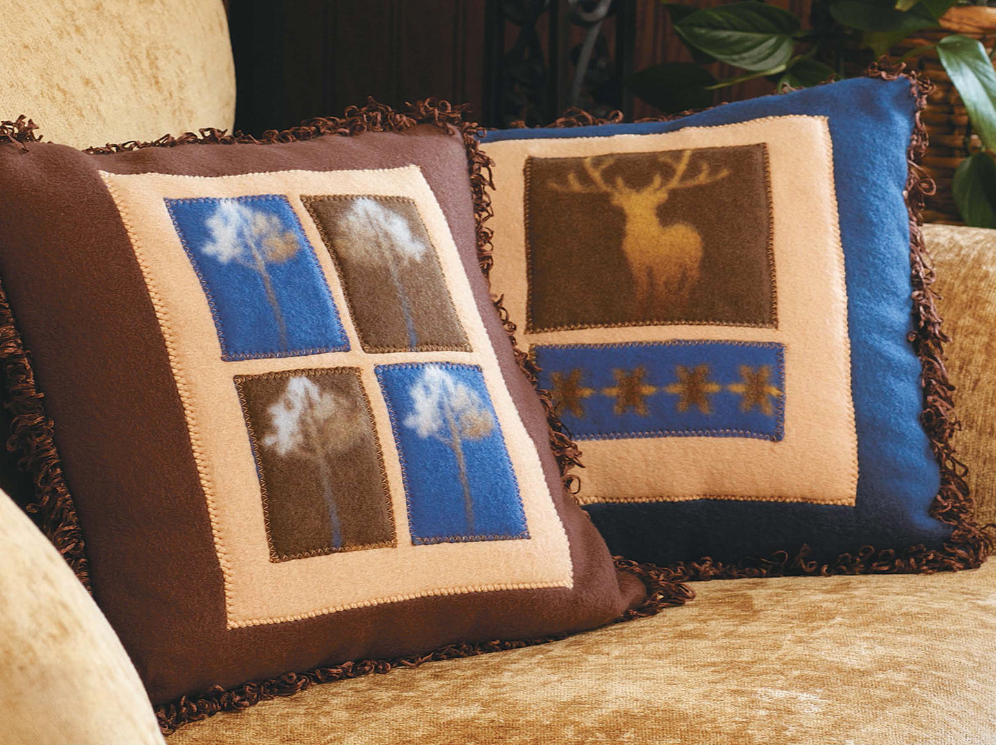 Two "Fleece at Home" pillows by Leisure Arts with rustic designs sit on a tan couch—one has trees in blue and brown squares, the other features a golden deer and stars. Both have brown fringe edges for extra charm.