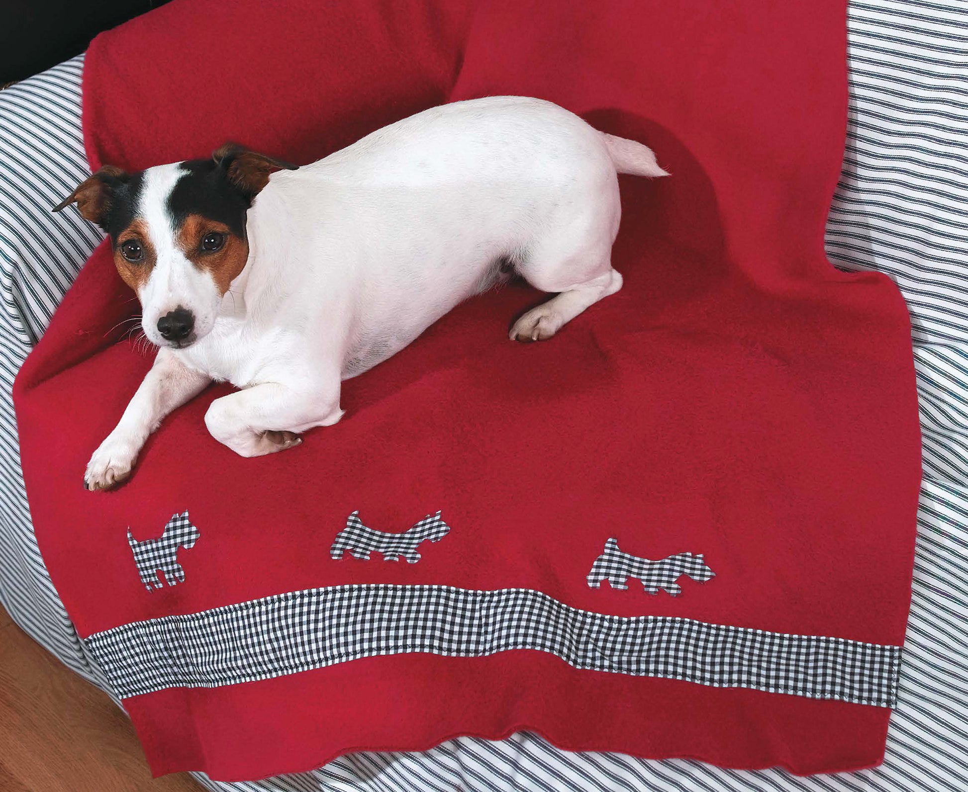 A small white and brown dog lies on the Leisure Arts Fleece at Home red blanket with plaid dog silhouettes, which is draped over a striped surface, possibly a bed with cozy pillows.