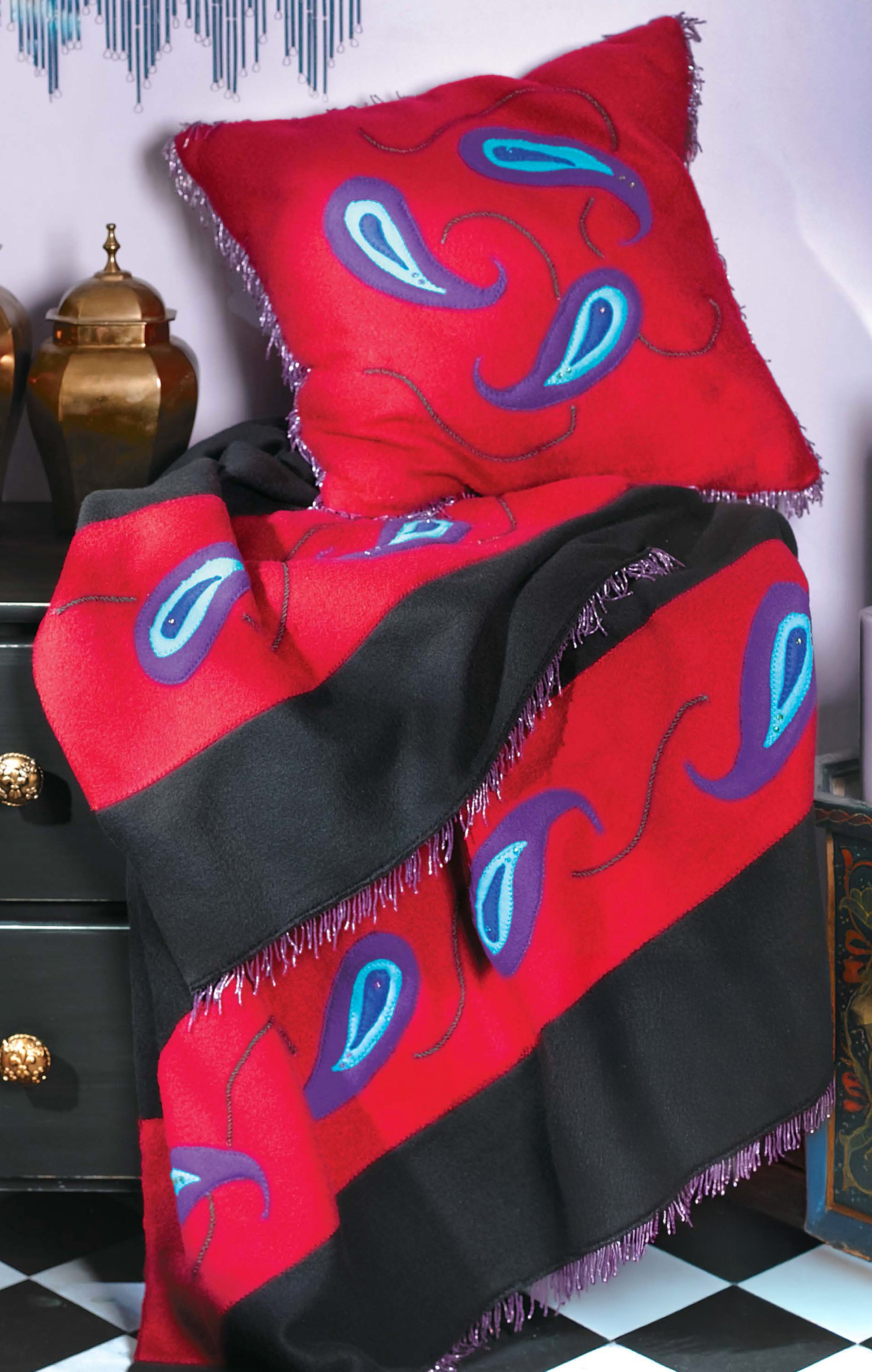 The Leisure Arts Fleece at Home throw blanket and matching pillows in red, black, blue, and purple paisley add stylish comfort, draped over a black chest with gold handles beside golden décor—perfect for easy throws.