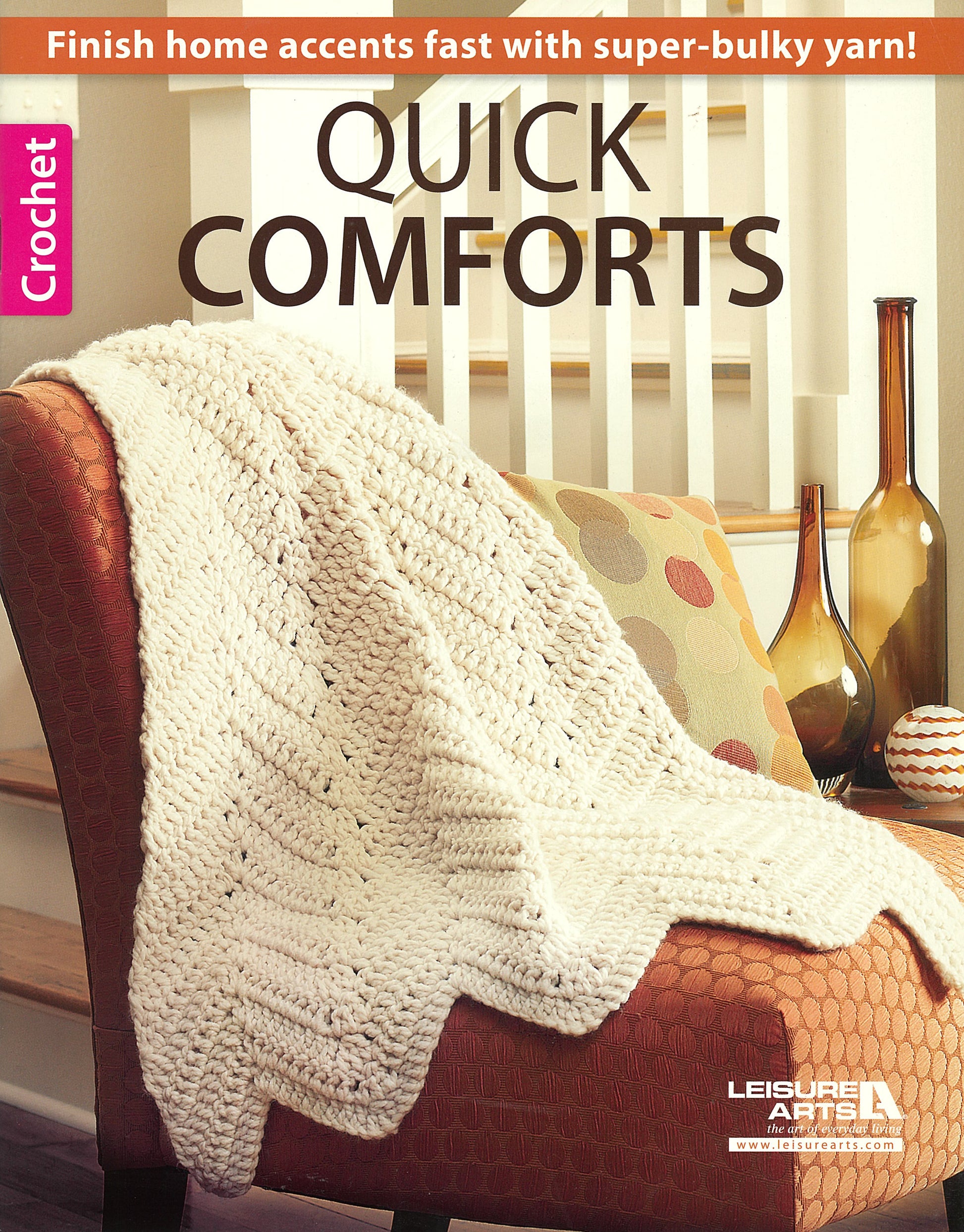 A cream-colored crocheted blanket drapes over a patterned armchair by a white railing. Text above: “Quick Comforts For Home by Leisure Arts—fast-finish afghans and home accents with super-bulky yarn!”.