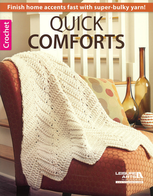 A cream-colored crocheted blanket drapes over a patterned armchair by a white railing. Text above: “Quick Comforts For Home by Leisure Arts—fast-finish afghans and home accents with super-bulky yarn!”.