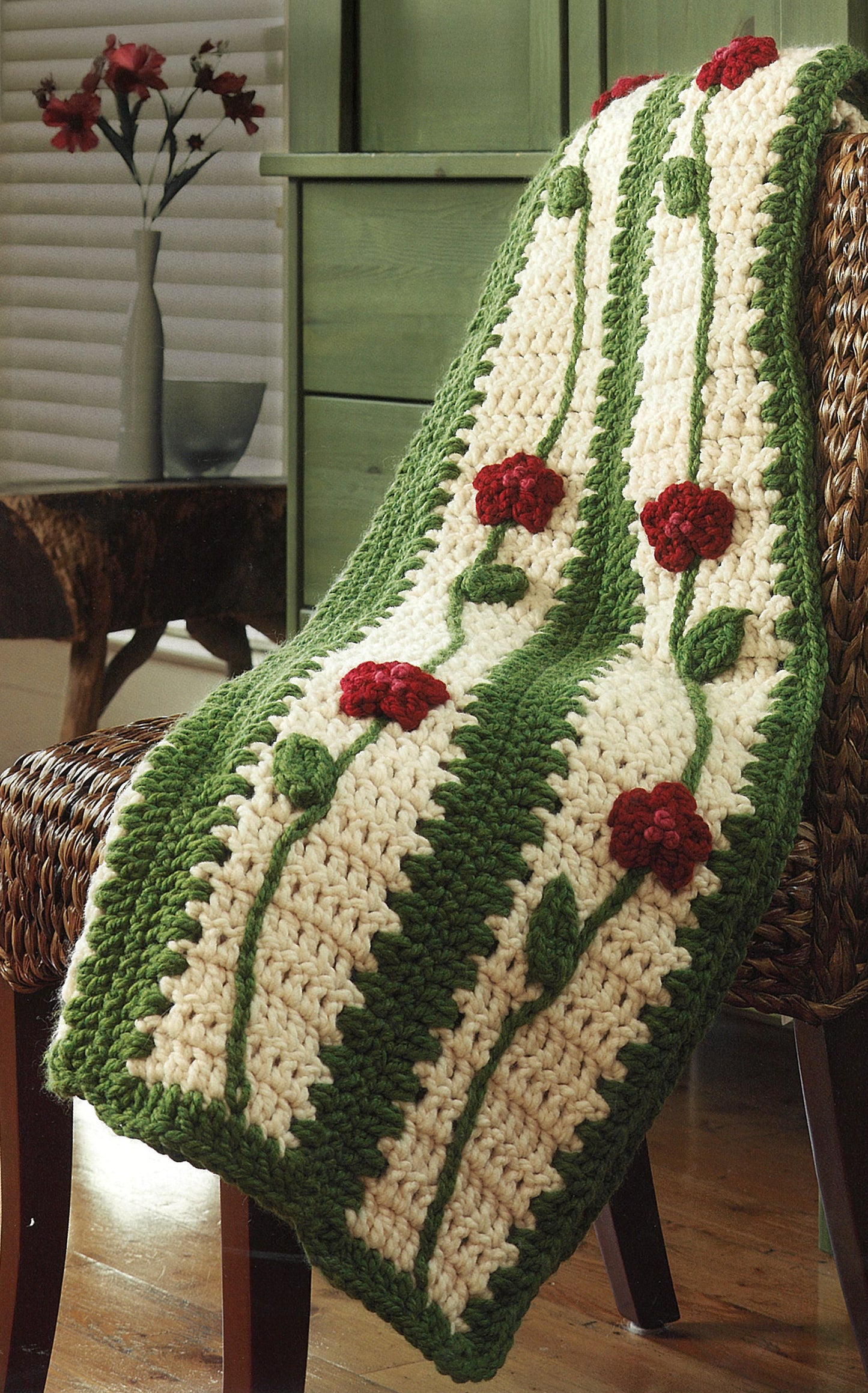 The Quick Comforts For Home crocheted blanket by Leisure Arts, made with super-bulky yarn, features green vertical stripes and red flower motifs, draped over a wicker chair; a vase of red flowers rests on a green dresser behind.