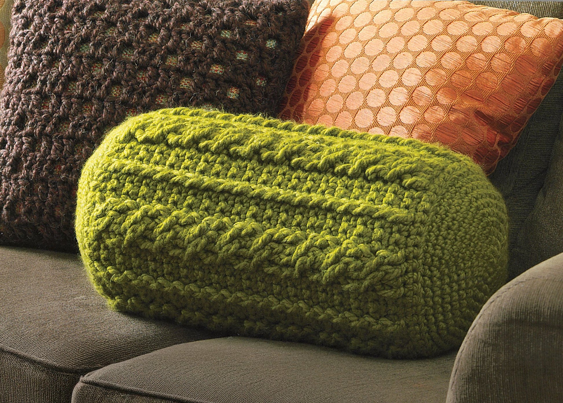 The Leisure Arts "Quick Comforts For Home" collection features a green textured cylindrical knit pillow paired with two square cushions—one orange with a circular pattern and one brown with chunky knit—adding warmth to any sofa.