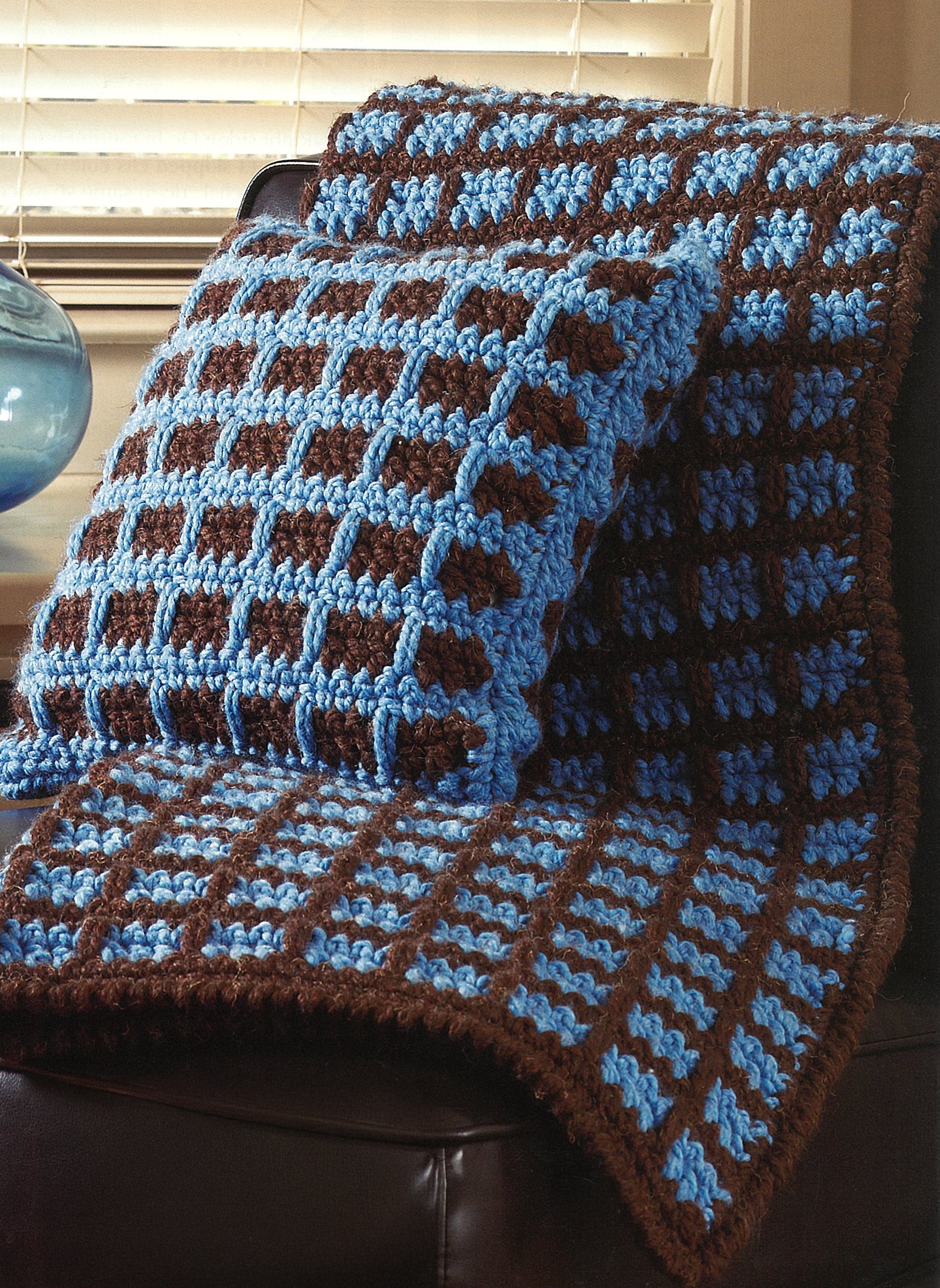 The Quick Comforts For Home set by Leisure Arts features a crocheted blue and brown checkered cushion and blanket made with super-bulky yarn, draped over a dark chair by a window with blinds.