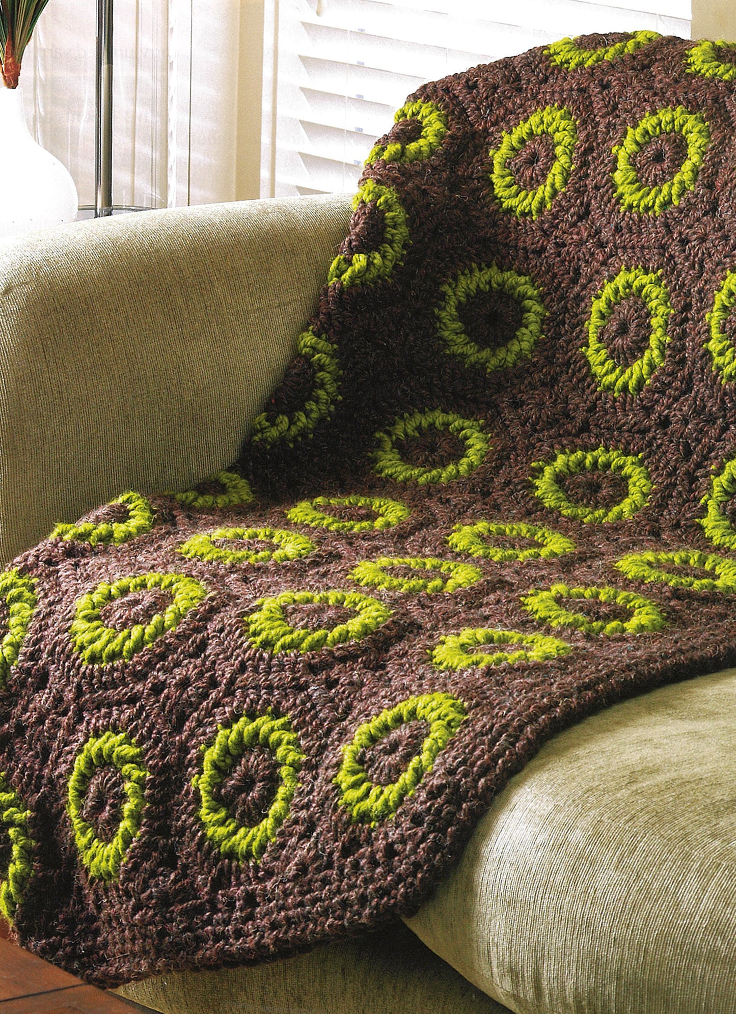 The "Quick Comforts For Home" brown crocheted blanket by Leisure Arts, featuring green circular rings in super-bulky yarn, is draped over a light-colored couch near a window with blinds.