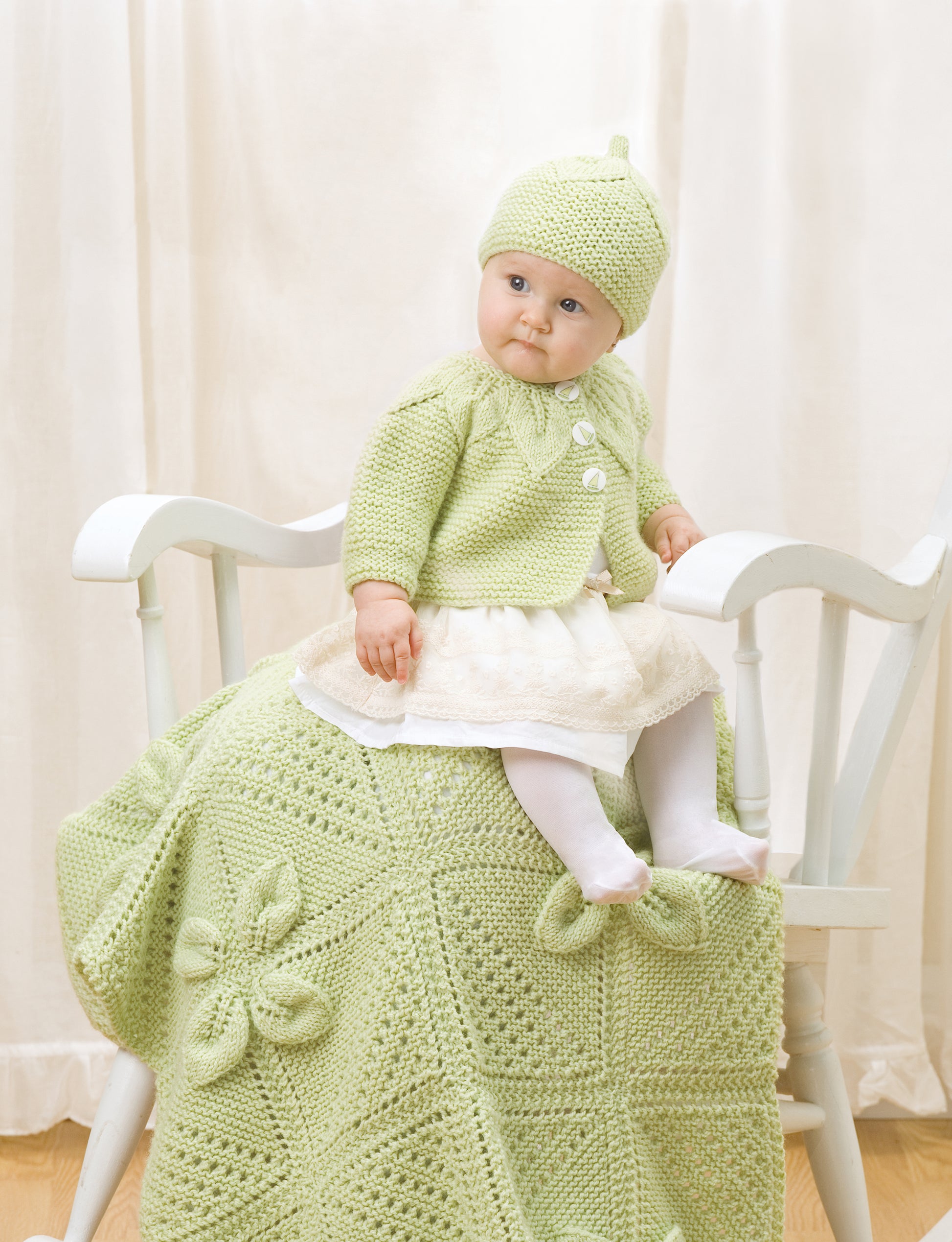 A baby models the Lace & Leaves for Baby set by Leisure Arts—hat, sweater, dress, and tights—seated on a flower-patterned crocheted blanket; ideal for those looking for intermediate knitting projects.