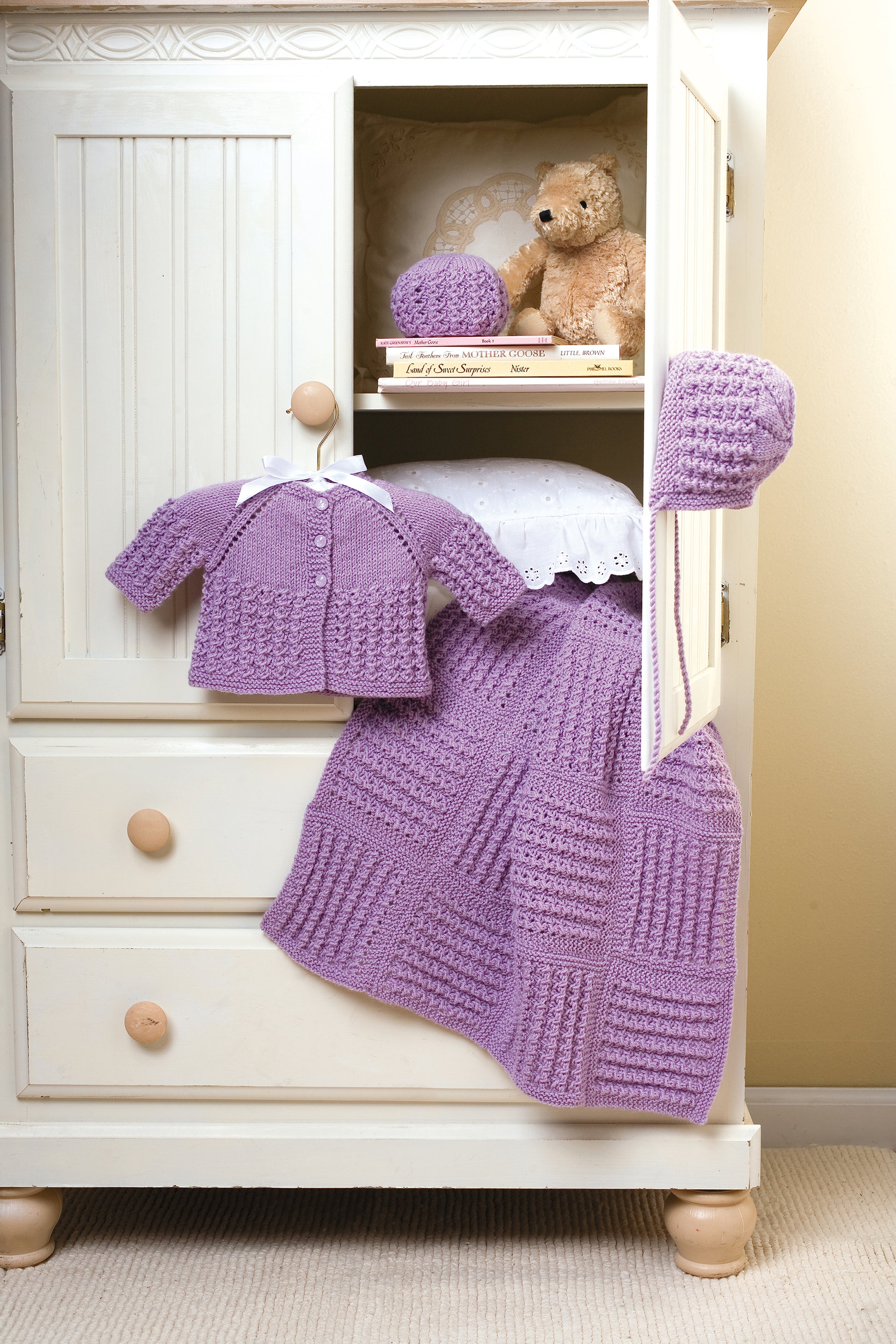 A cream cabinet with an open door showcases the Leisure Arts "Lace & Leaves for Baby" set: a matching lavender knit sweater, hat, and blanket. A teddy bear, doily, and book add to the cozy, neat display.