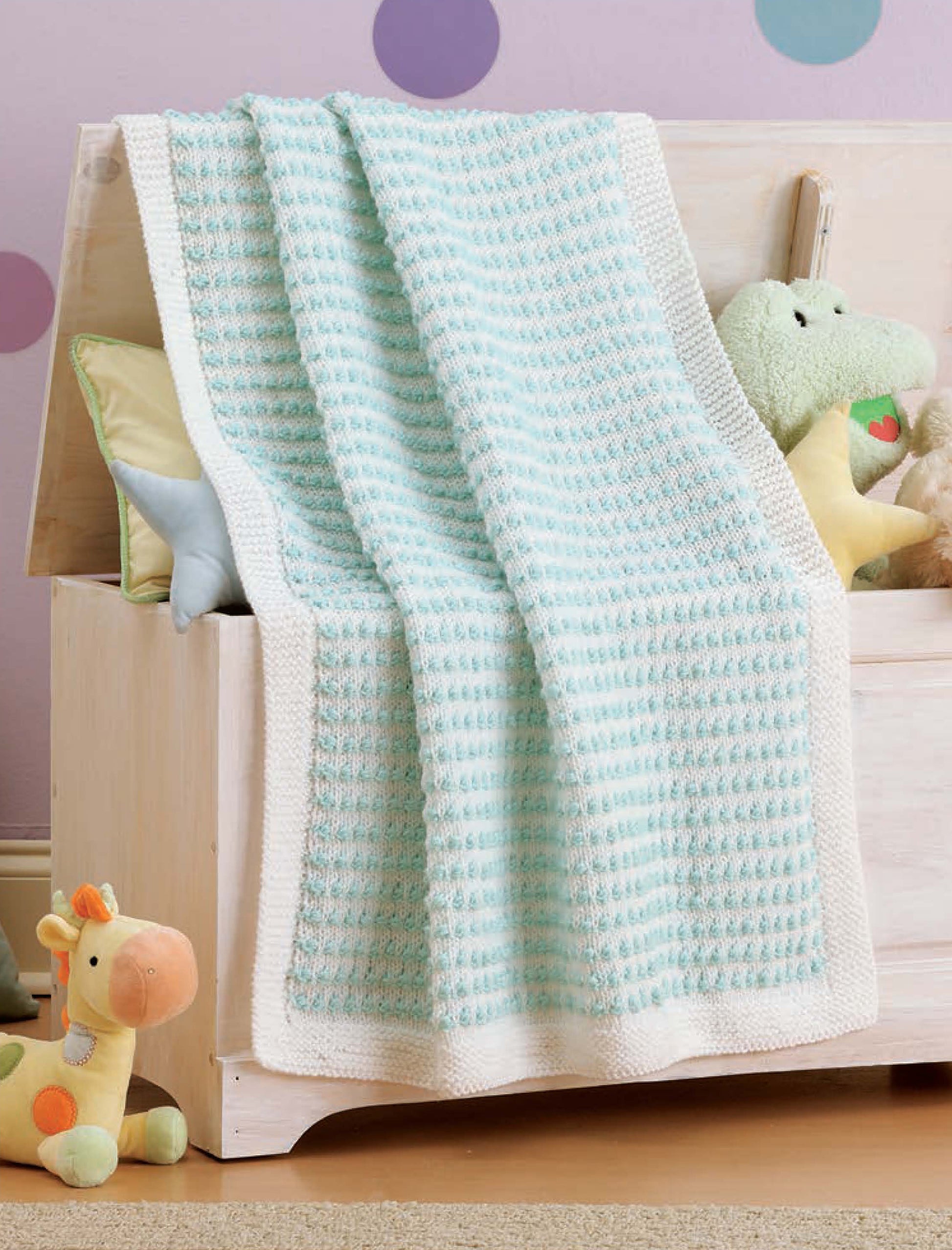 A baby blanket made from Leisure Arts’ Little Lullabies—5 Knit Baby Afghans pattern in light blue and white rests on a wooden chest, surrounded by pastel pillows and plush toys in a nursery with purple polka dot walls.