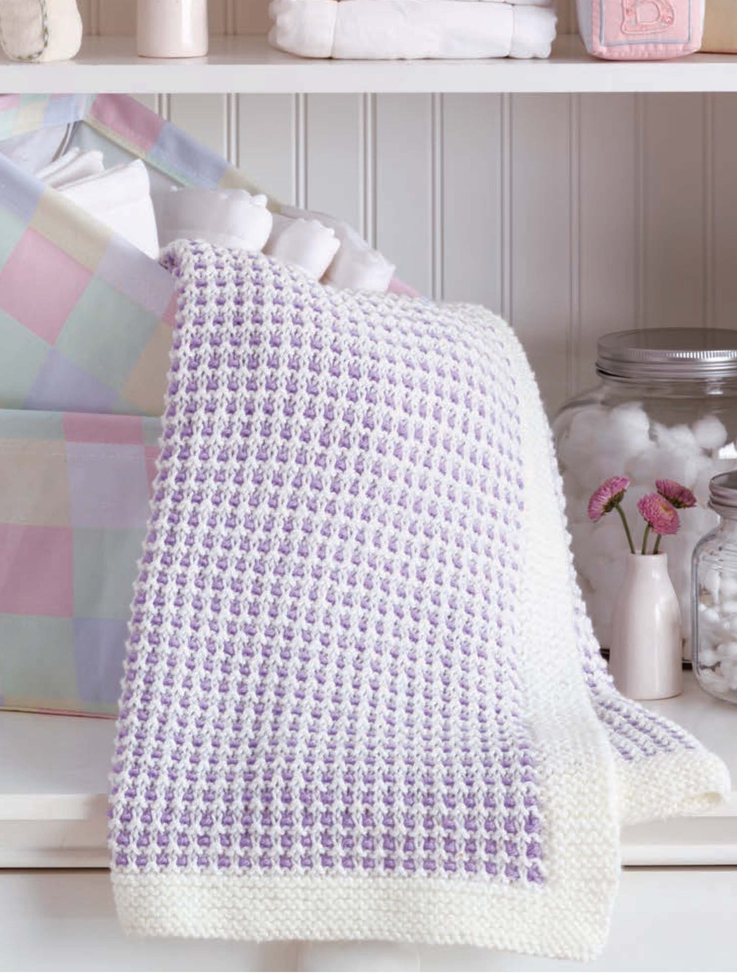 The Little Lullabies—5 Knit Baby Afghans by Leisure Arts, featuring a white and lavender checkerboard pattern, is draped over a pastel storage basket with shelves behind holding towels, jars, and pink flowers.