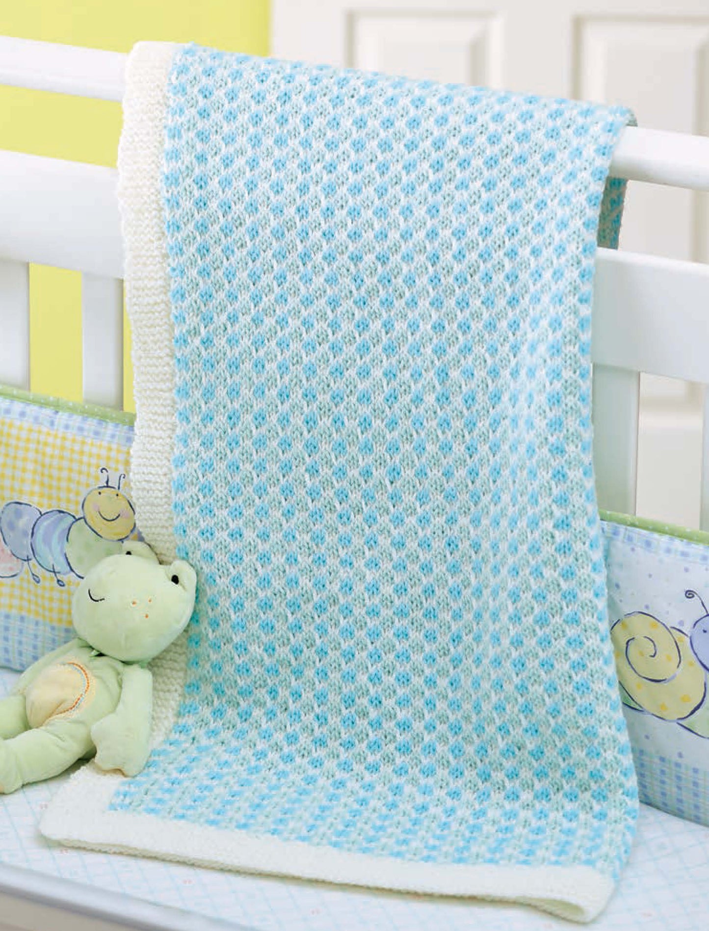 A soft blue and white blanket from Little Lullabies—5 Knit Baby Afghans by Leisure Arts hangs on a crib rail. A green frog toy is nearby, and the bumper showcases playful caterpillar and snail designs. Perfect for baby blanket knitting fans!.