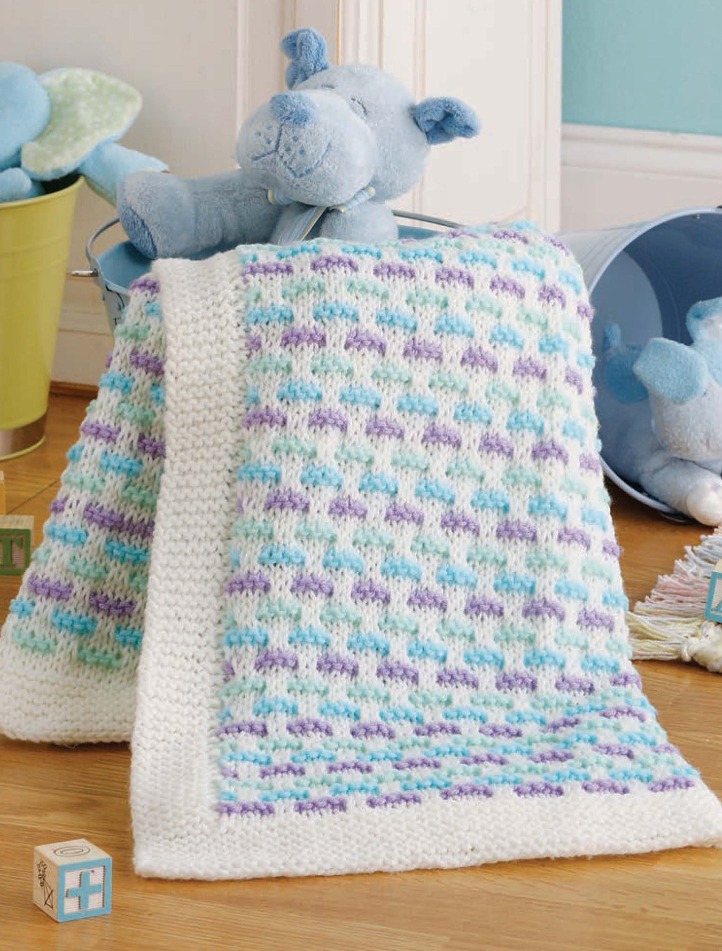 Leisure Arts’ Little Lullabies—5 Knit Baby Afghans features a soft, handmade baby blanket with a white border and blue, purple, and teal patterns, draped over a basket near a blue stuffed bear and toys on a wooden floor.