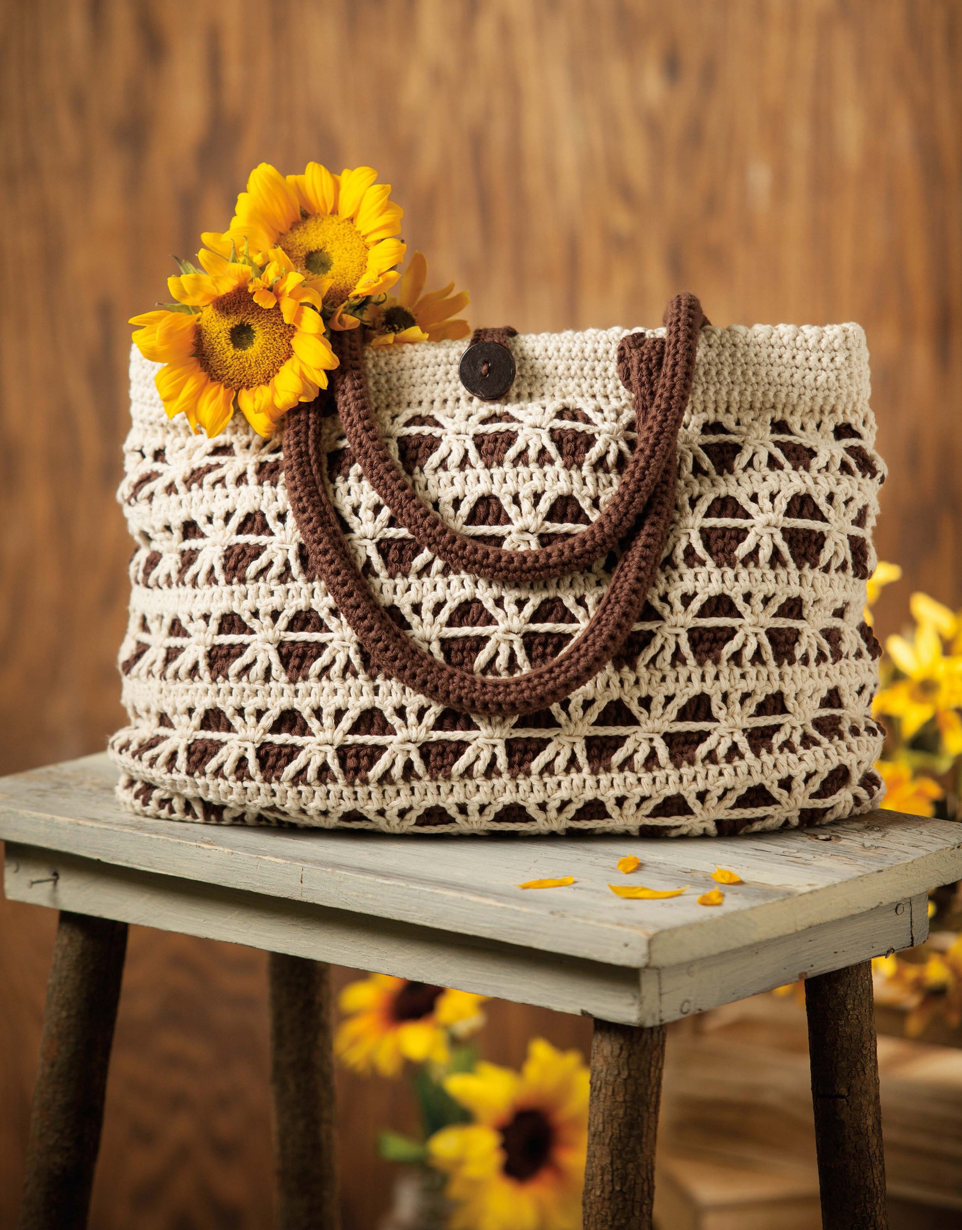 The Crochet Terrific Totes—Dress to Impress With Distinctive Bags! digital download from Leisure Arts is featured here: a beige and brown crocheted tote sits on a stool, filled with bright yellow sunflowers, with more flowers in the background.