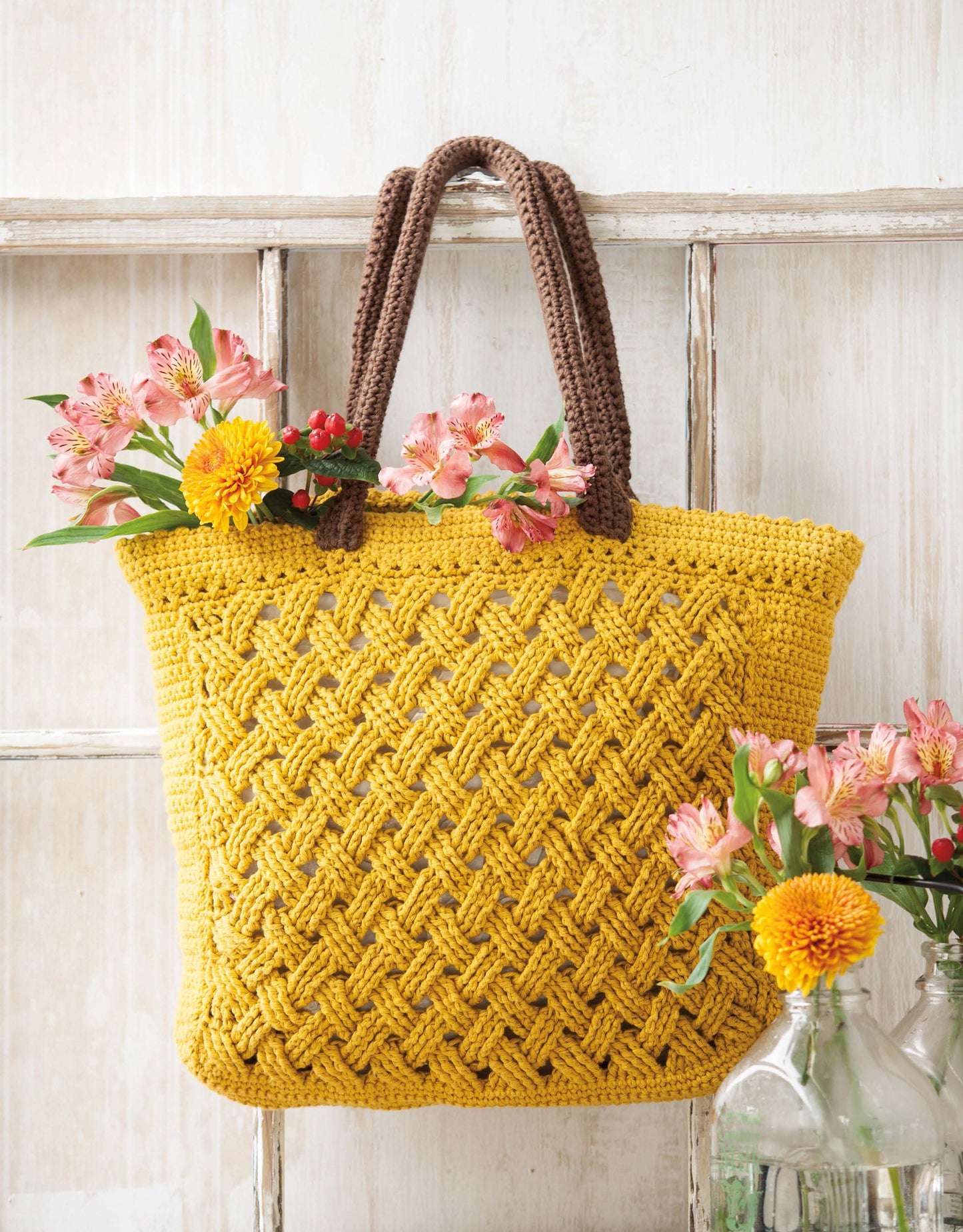 The Crochet Terrific Totes—Dress to Impress With Distinctive Bags! Digital Download by Leisure Arts is displayed on a rustic window frame with colorful spring flowers and bottles, adding cheerful charm to any setting.