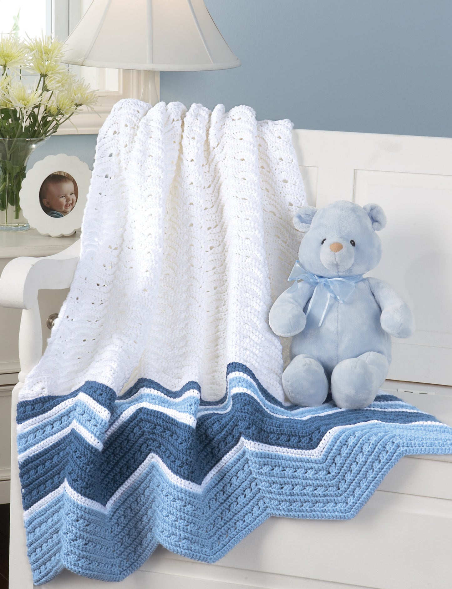 The Terrific-to-Touch Baby Afghans by Leisure Arts, featuring white and blue nursery crochet patterns, drapes over a dresser beside a light blue teddy bear. A lamp, flowers, and baby photo decorate the softly lit room.