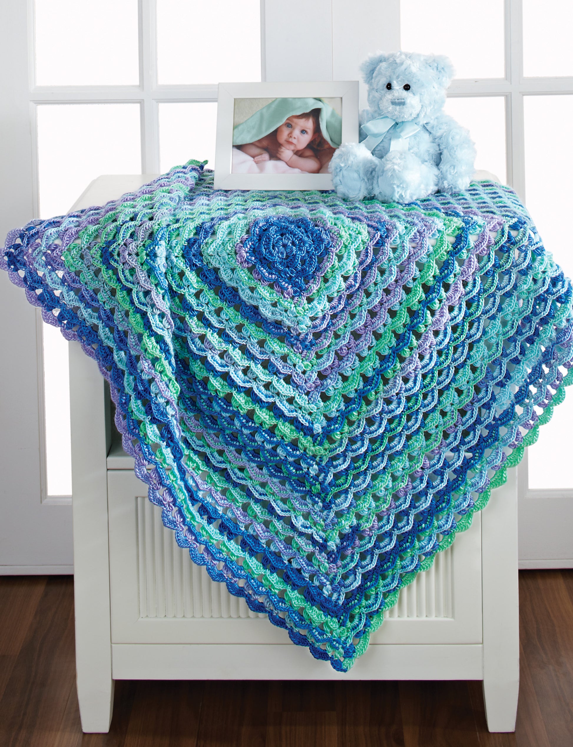 A vibrant afghan from *Terrific-to-Touch Baby Afghans—The Best of Mary Maxim* by Leisure Arts in blue, teal, and purple lies on a white chest with a blue teddy bear and baby photo. White French doors stand in the background.