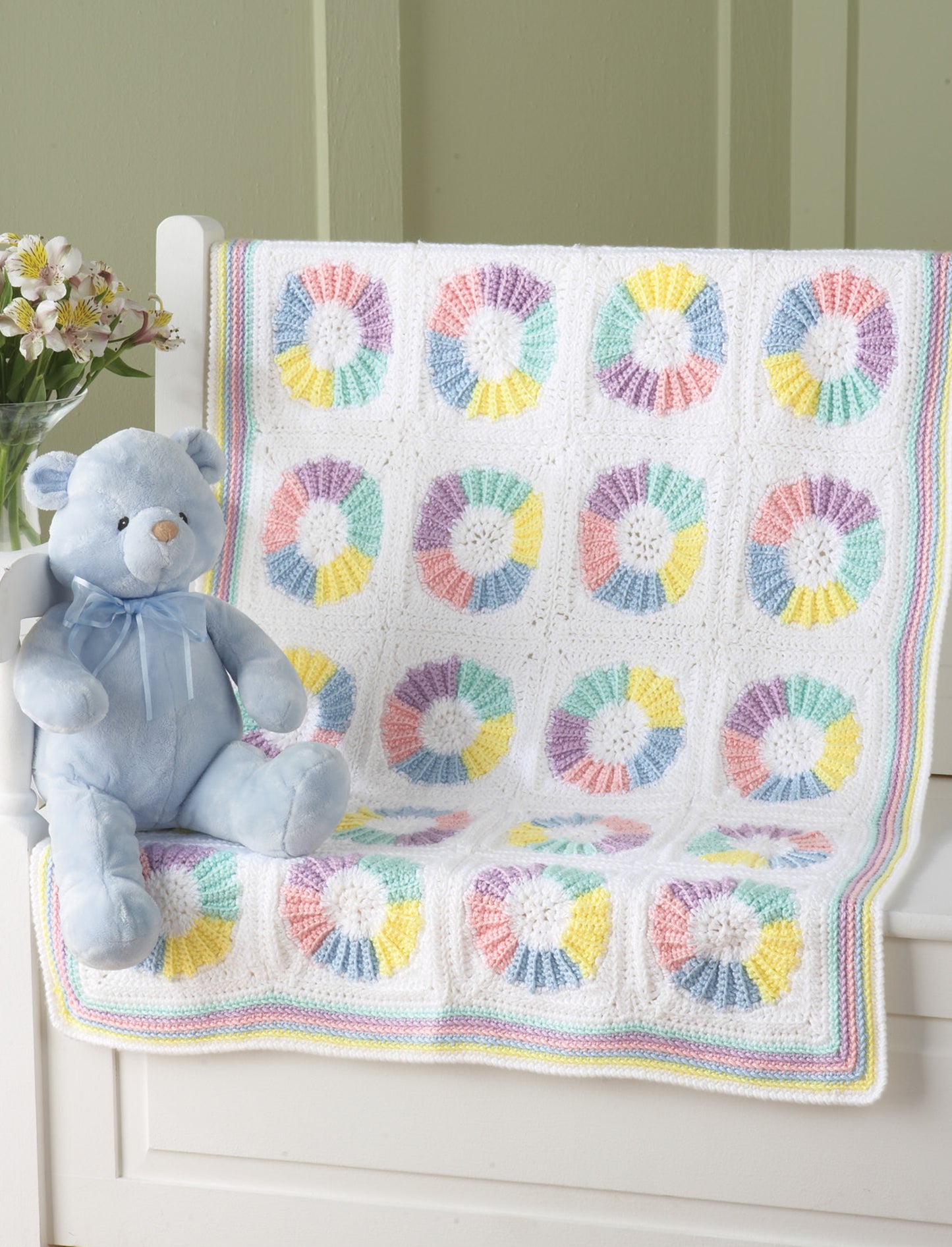 The Terrific-to-Touch Baby Afghans from Leisure Arts, featuring pastel patchwork and circular designs, is draped over a white bench next to a light blue teddy bear, with a vase of light flowers placed behind.