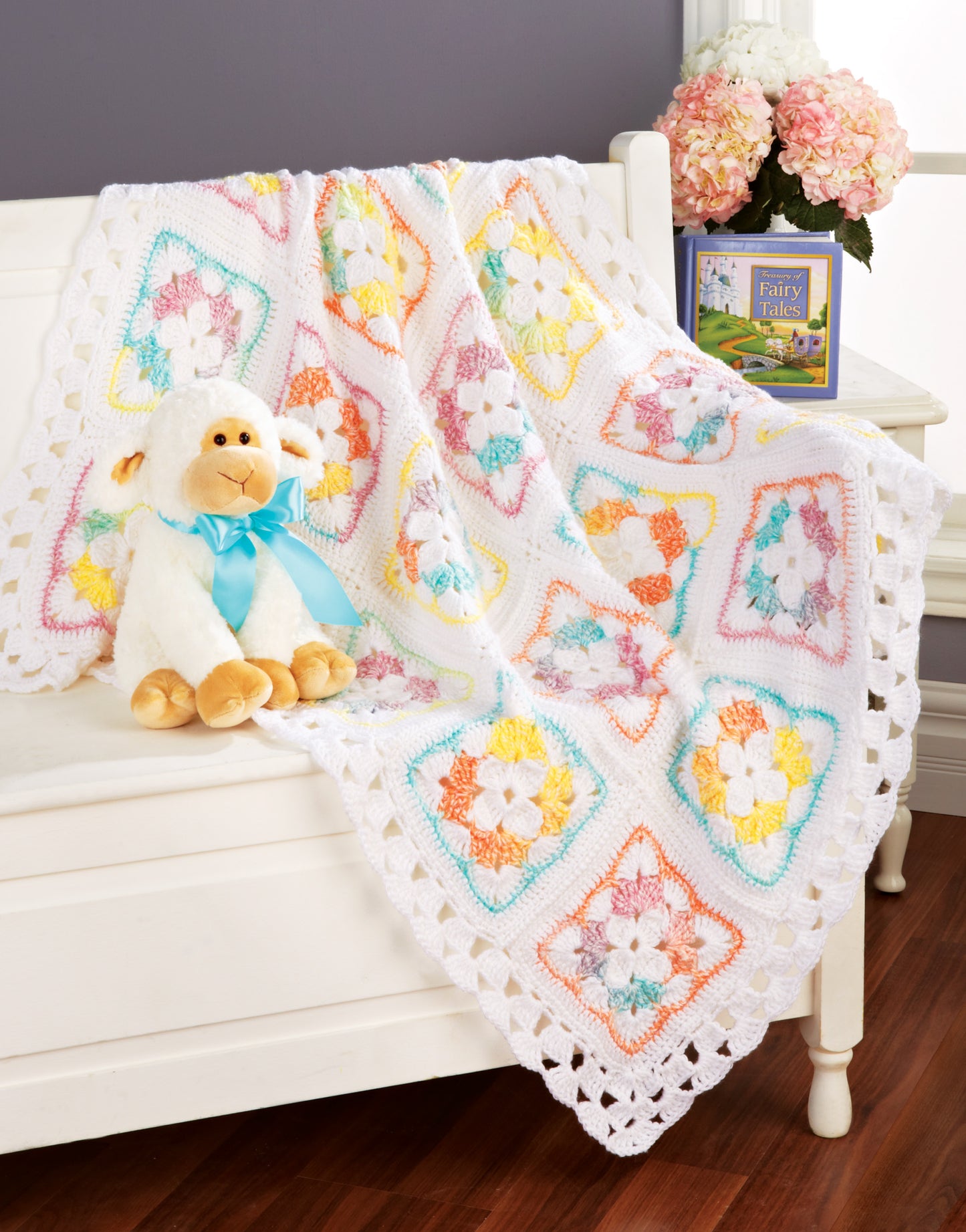A plush lamb toy with a blue ribbon sits on a white bench draped with the "Terrific-to-Touch Baby Afghans—The Best of Mary Maxim" by Leisure Arts; behind it are a book and vase of pink flowers, set against a gray wall and wooden floor.