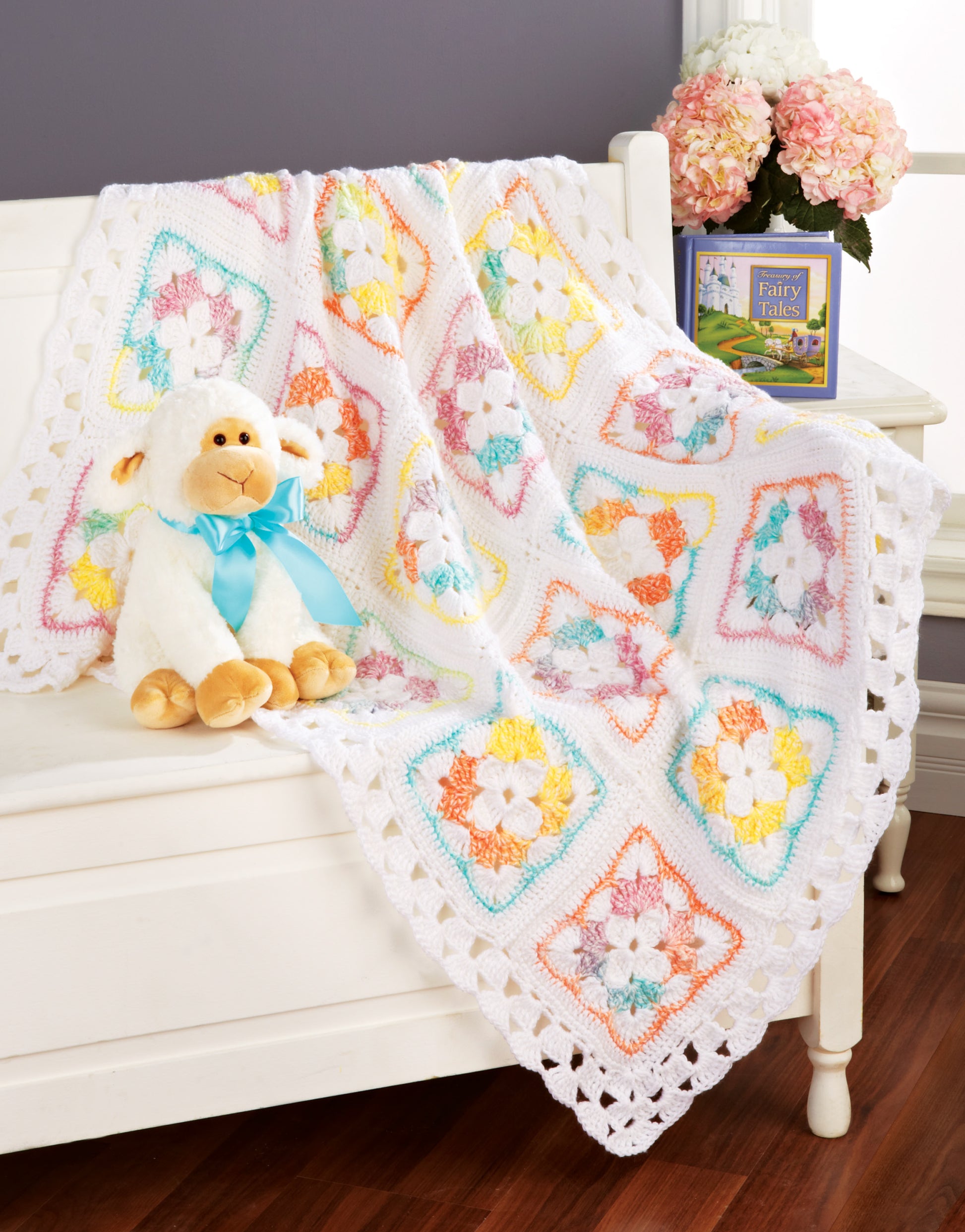 A plush lamb toy with a blue ribbon sits on a white bench draped with the "Terrific-to-Touch Baby Afghans—The Best of Mary Maxim" by Leisure Arts; behind it are a book and vase of pink flowers, set against a gray wall and wooden floor.