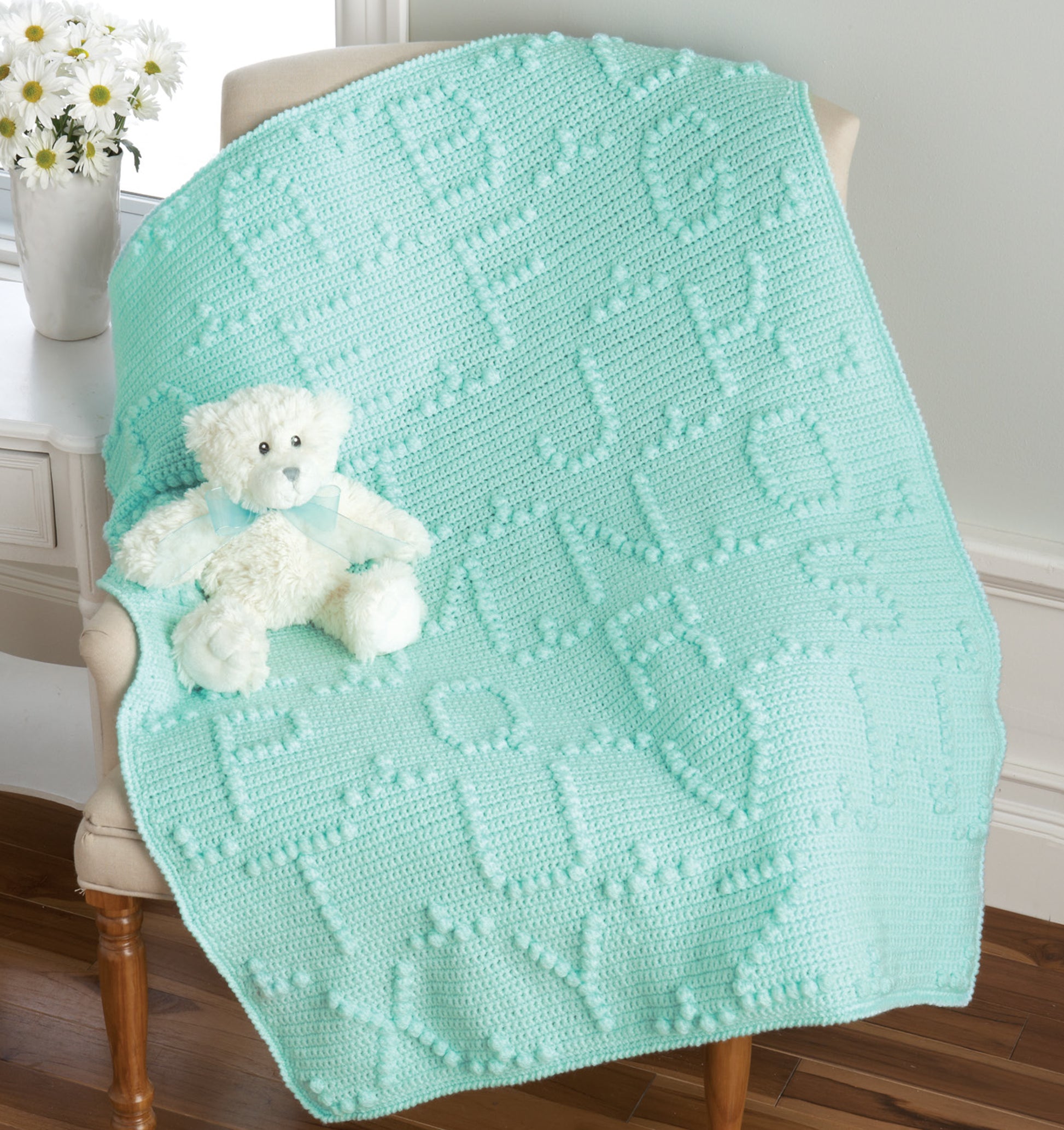 The Terrific-to-Touch Baby Afghans from Leisure Arts, in refreshing mint green with raised alphabet letters, is draped over a white chair topped with a white teddy bear. A vase of daisies sits on the windowsill nearby.